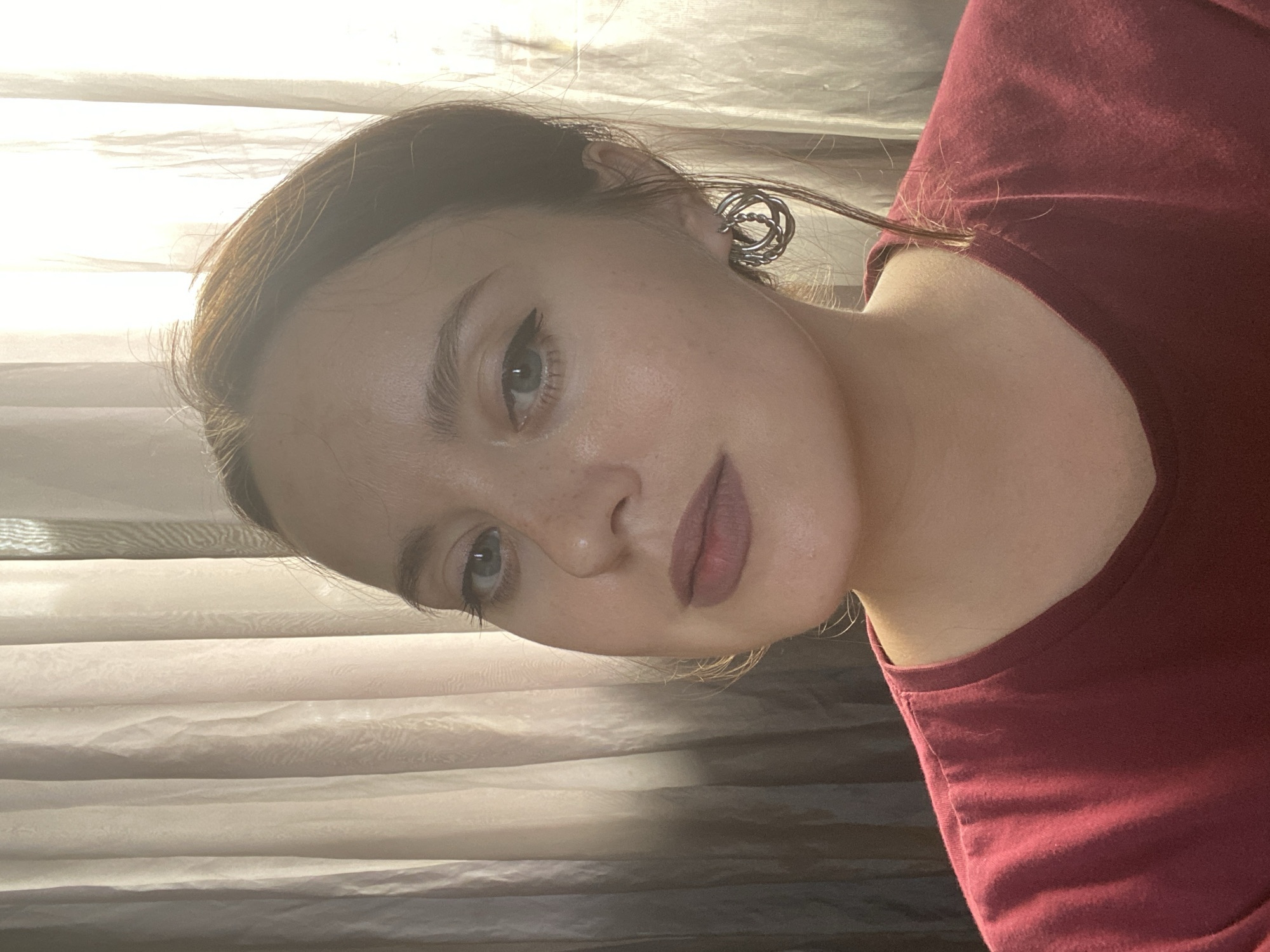 The image shows a close-up portrait of a young Caucasian woman with light skin, freckles, light brown hair pulled back, and green eyes. She is wearing a maroon t-shirt and large silver hoop earrings. The background consists of sheer, vertically draped curtains. The lighting suggests natural light, possibly from a window, casting shadows on the curtains and illuminating the woman's face from the side. There are no identifiable elements in the image that would allow for determining a specific location within the city of Batumi, Georgia.