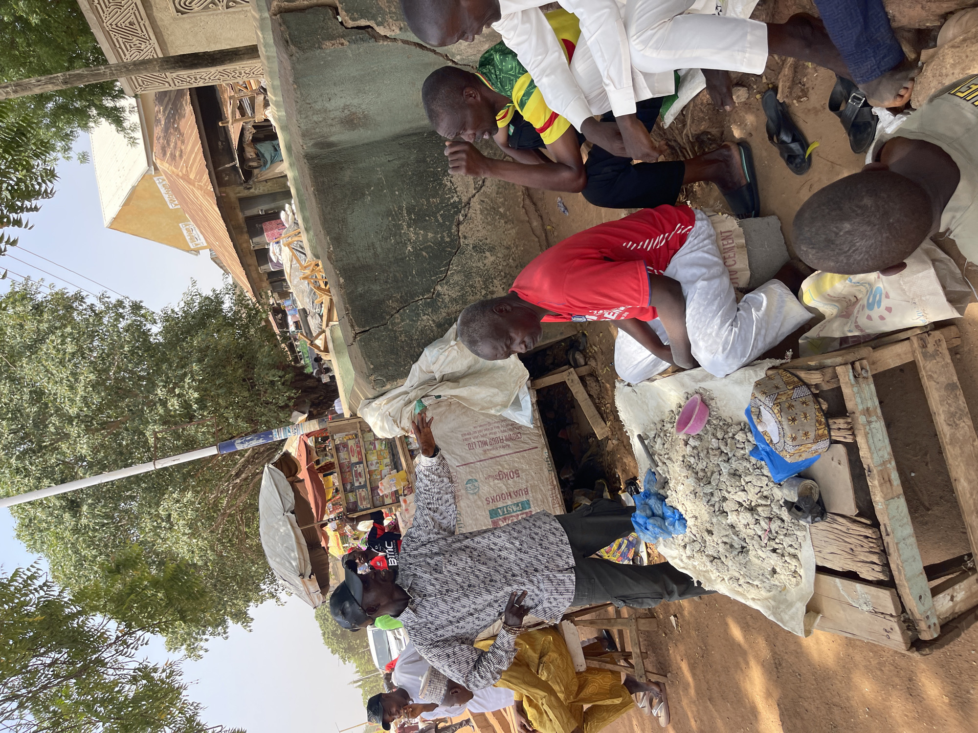 This outdoor market scene captures a group of men engaged in commerce and daily activities, likely in Zaria, Nigeria. The image is taken from a slightly elevated perspective, looking down into a bustling area with various stalls and goods.
In the foreground, several men are seated or kneeling on the dusty ground. One man, wearing a red shirt and white trousers, crouches next to a white sack, seemingly attending to items. Beside him, another man in a yellow and green patterned shirt sits, hands clasped. Other men in white clothing are also seated nearby. Their footwear ranges from sandals to slippers.
The central focus of the market activity involves a wooden stall or makeshift table covered with a white cloth, displaying a large pile of irregular, light-colored, granular lumps—possibly a local food item or commodity. A small pink bowl and a patterned bag are also on this display. Nearby, a standing man in a black and white patterned shirt and dark cap is handling a large white sack, which appears to be a major part of the trade.
Visible text on one of the large white sacks reads "CROWN FLOUR MILL LTD", "50KG", "PASTA", and "BUJA HOOKS", indicating the sale of flour or related food products.
The background features more market stalls with shelves stocked with various packaged goods. An open umbrella provides shade. Beyond the market area, dense green trees line the street, and several buildings are visible, including one with an ornate, carved facade, and others with simpler, possibly corrugated iron roofs. The ground is a reddish-brown, dusty earth, consistent with an outdoor market environment. The bright natural light indicates it is daytime, with clear weather. The scene depicts a lively but calm atmosphere of daily life and trade.