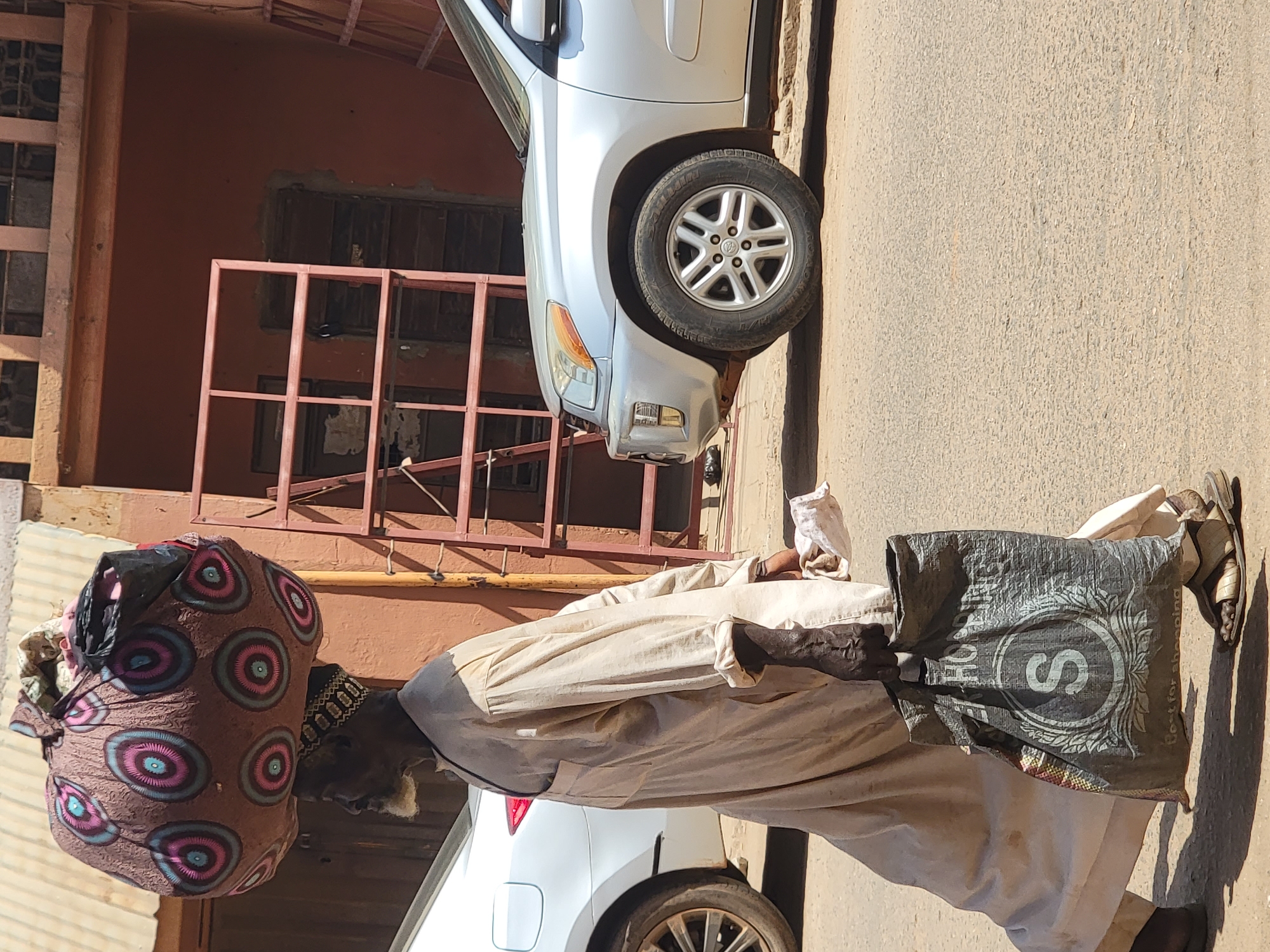 In an outdoor setting in Maiburiji, Nigeria, a person is walking along a street adjacent to parked vehicles and buildings. The person carries a large, patterned bundle on their head and a shopping bag in their hand. The bag has a prominent "S" on it, surrounded by a decorative design. The person's attire includes a light-colored, flowing garment and sandals. The street is paved and shows signs of wear. A white car is parked nearby. The buildings feature windows and architectural details.