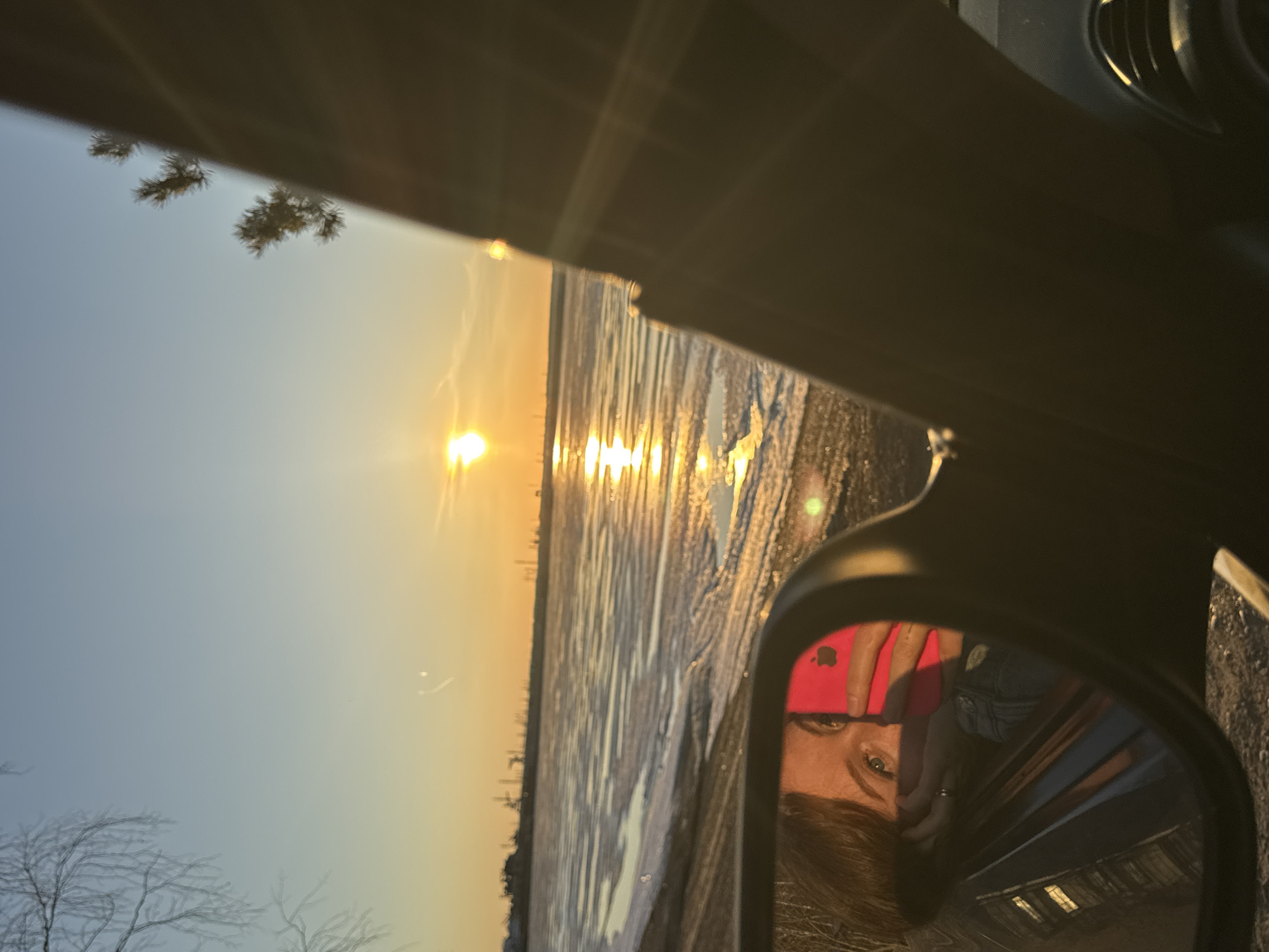 The image shows a sunset reflecting on a lake or river, with a person's reflection visible in a car's side mirror. The person appears to be holding a phone. The location is identified as Chaikovsky, Russia based on the landscape and architectural details. The environment is cold, potentially winter-like, with ice or snow on the river.