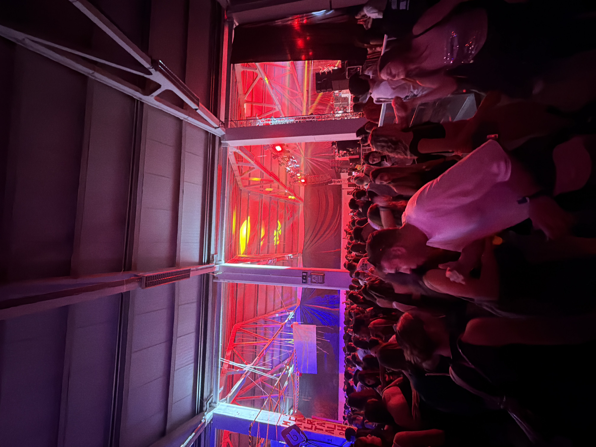 The image captures an indoor event, likely a concert or performance, taken from an elevated side perspective. The scene is rotated 90 degrees clockwise.
**Subjects:** A large audience occupies the right side of the frame, consisting of many people standing and facing towards a stage or performance area to their left. Individuals in the crowd are dressed casually, with some faces partially illuminated by the ambient lighting. The left side and upper portion of the image reveal the venue's architecture, characterized by prominent white metal trusses and structural beams that form part of the building or stage setup. Various stage lighting fixtures, emitting vivid red and yellow light, are mounted on these structures. In the midground, a stage area is visible, complete with additional lighting rigs and a large banner or screen displaying a blue and white design. The left wall of the venue appears to be constructed from paneled or corrugated material, bathed in a soft purple light.
**Activity or event taking place:** The image clearly depicts an active indoor music concert, performance, or large social gathering. The crowd's orientation suggests they are engaged in watching an event on the stage.
**Notable details:** The event is taking place at night, illuminated entirely by artificial light. The dominant color palette of the lighting is a striking combination of red and yellow, creating an energetic and immersive atmosphere. The presence of extensive structural elements and a sizable crowd indicates a large-scale venue. The location is Bragança, Portugal.
**Visible text:** A small, white rectangular sign with the number "16" in red text is visible on a barrier or structure positioned in front of the crowd, near the bottom center of the image. Below the number "16", partially obscured text appears to read "BRAGA...".