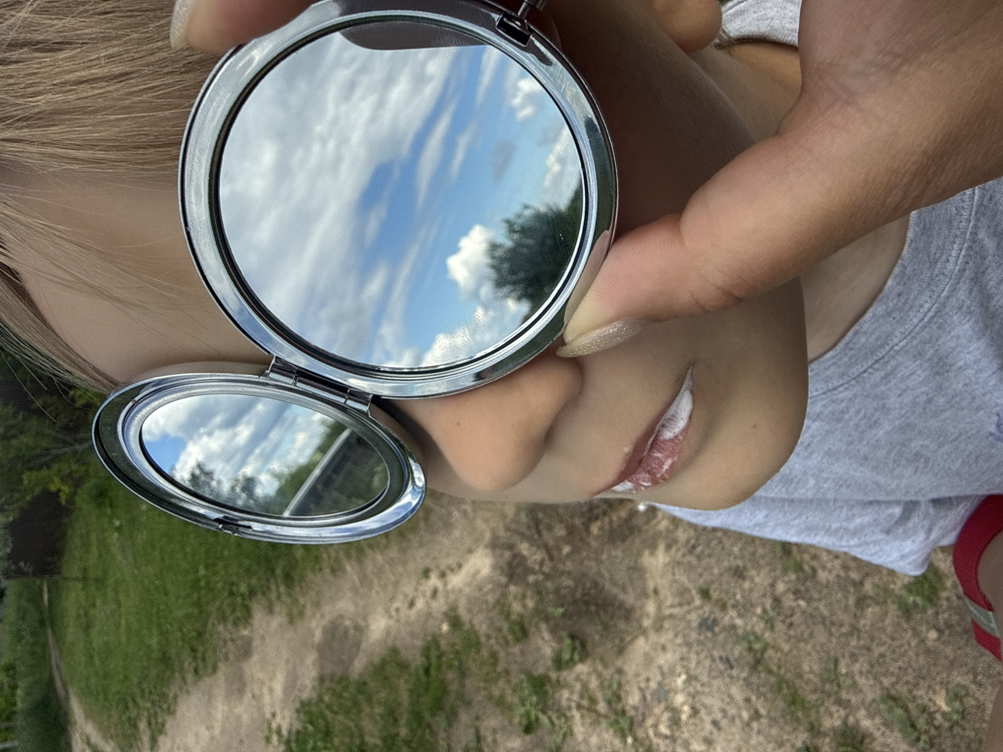 In this close-up, vertically oriented photograph, a person's face and hands are visible as they hold a compact mirror. The mirror reflects a blue sky with fluffy white clouds and a hint of green foliage, suggesting an outdoor setting on a clear day. The person's fair hair is visible at the top of the frame, and their lips, adorned with what appears to be white lipstick or balm, are a focal point. One hand, with manicured nails, is holding the top part of the mirror, while another hand holds the bottom, open part, which also reflects the sky and some surrounding greenery. The background of the image shows a path or ground covered with dirt and sparse vegetation, and in the distance, more greenery. The overall impression is that of a personal moment, possibly taken outdoors in a natural environment. There is no visible text in the image. The location context is Dmitrov, Russia.