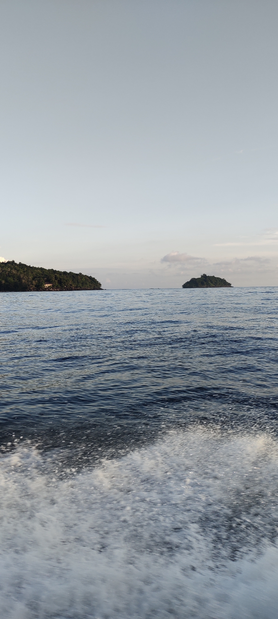 The image shows a view of the sea with a coastline and islands in the distance. In the foreground, a boat's wake is visible, creating white foam on the dark blue water. On the left side of the frame, a lush green coastline stretches out, with a small cluster of buildings visible among the trees. Further out in the sea, a small, uninhabited island covered in dense green vegetation stands isolated. The sky above is a soft, pale blue with a few wispy clouds, suggesting it is either early morning or late afternoon. The overall scene depicts a tranquil tropical seascape. This scene is likely located in the waters surrounding Sabang, Indonesia, an archipelago known for its beautiful islands and clear seas.