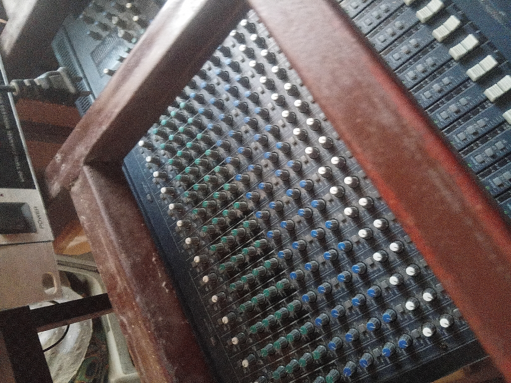 This image, captured in Jalingo, Nigeria, displays an audio mixing console set within a rustic, reddish-brown wooden frame. The main mixing board, dark grey in color, occupies the central and left portions of the frame, showing numerous rotary knobs arranged in rows. These include a top row of white knobs, multiple intermediate rows of green knobs, and several lower rows of blue knobs. The surface of the mixer exhibits a noticeable layer of dust. In the lower right quadrant, partially obscured by the wooden frame, another section of a mixing console is visible, featuring multiple linear faders (sliders). In the upper background, behind the wooden frame, a white electronic device marked "POWER" is visible, along with a black cable. To the upper right, another audio mixing console is partially visible, also featuring rows of knobs. No individuals are present in the scene.