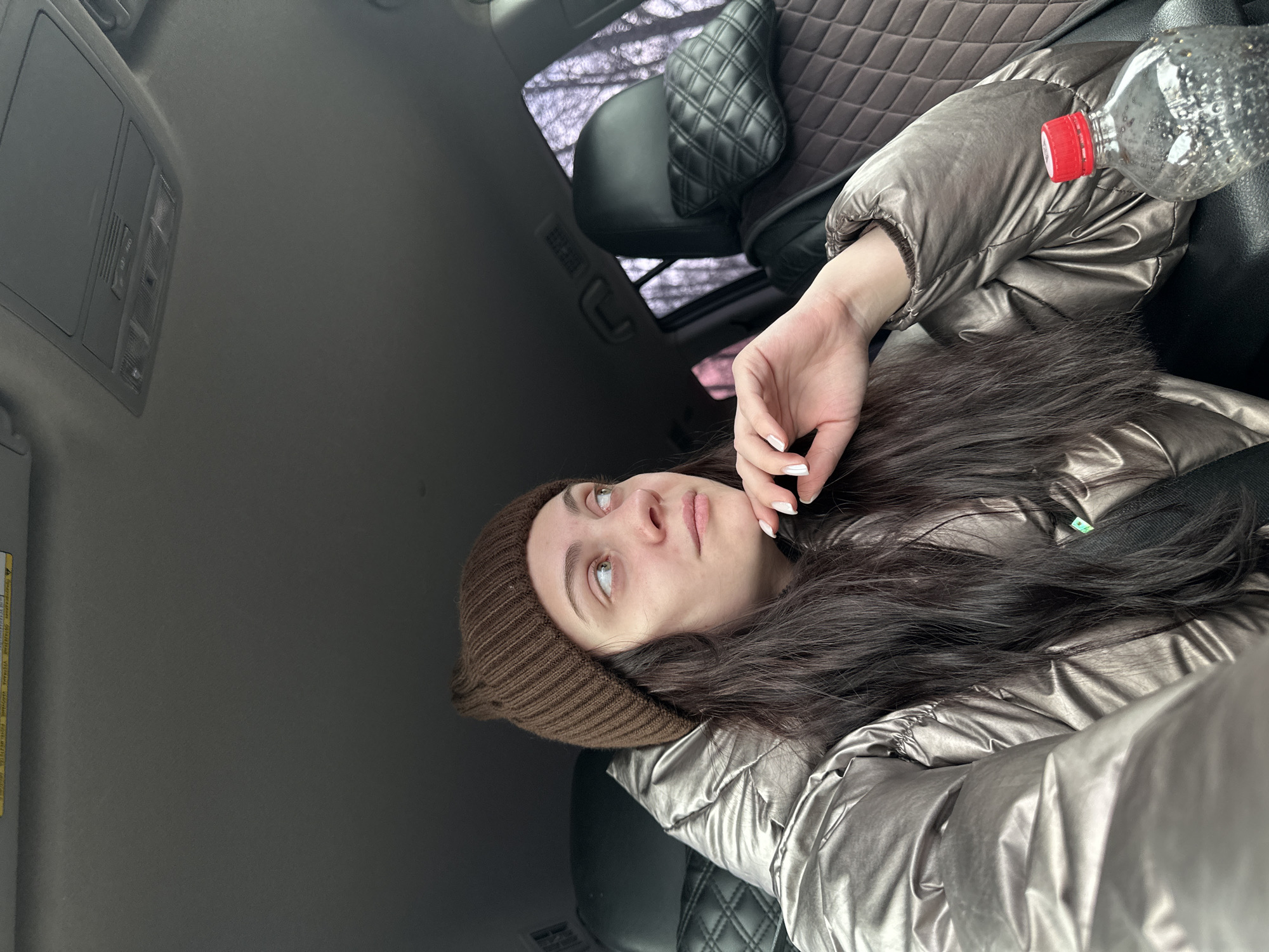 A young woman wearing a brown beanie and a shiny brown puffer jacket is reclining in a car. Her long, dark hair is draped across her jacket. Her left hand is raised near her face, and a water bottle with a red cap sits beside her on the seat. The car's interior is gray, with a black quilted headrest visible in the background. The outside scenery, visible through the car window, consists of bare trees, but location cannot be confirmed.