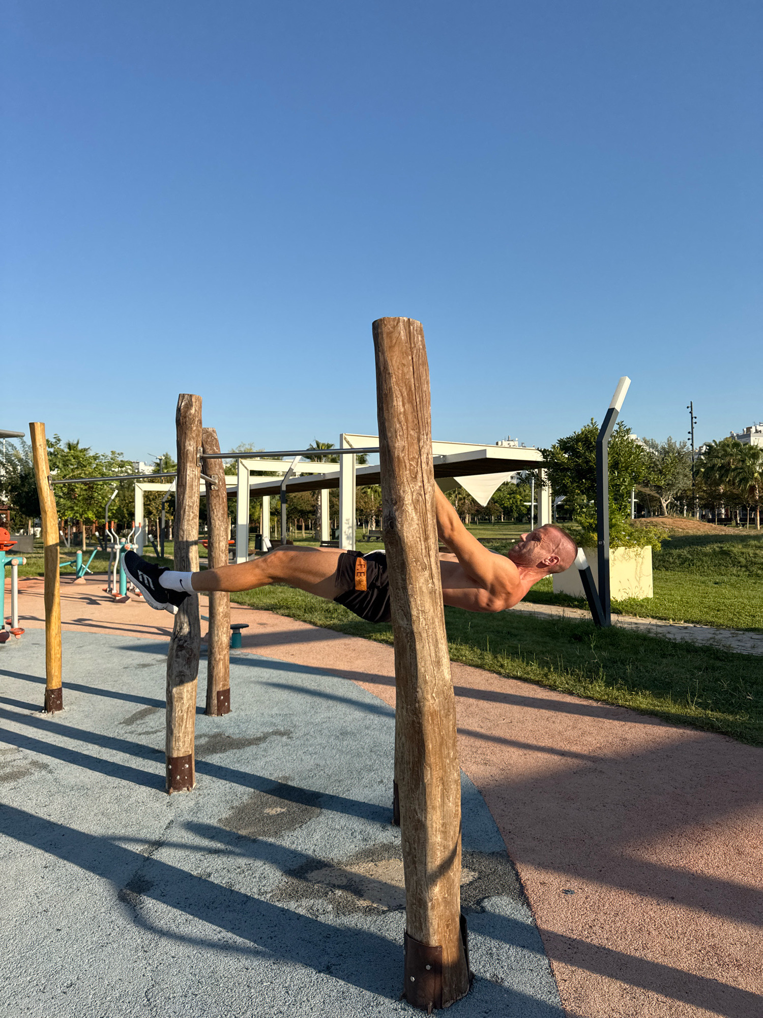 A bald, muscular man is performing a front lever exercise on wooden bars in a park in Antalya, Turkey. He is shirtless and wearing black shorts with an orange design. His body is horizontal to the ground, supported by his arms. The park has a blue and orange rubberized ground, with grass and trees in the background. There are other exercise machines visible in the distance, and the sky is clear and blue. The sun is casting long shadows from the wooden bars.