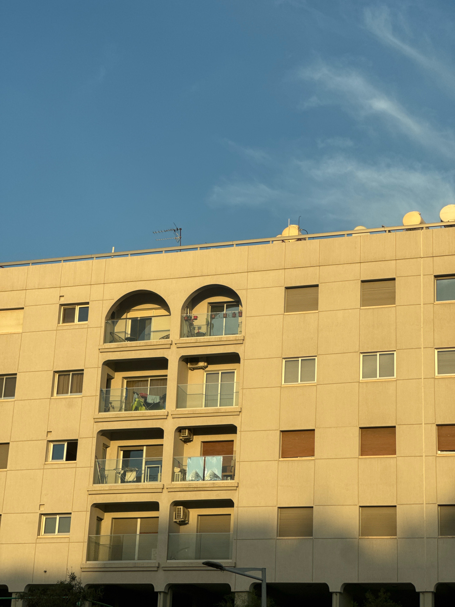 The image shows a portion of a pale yellow, multi-story apartment building under a clear blue sky with some wispy white clouds. The building has a facade of square panels, with numerous windows and several balconies. The windows are mostly rectangular, some with horizontal blinds, and others with glass panes. The balconies are recessed and feature glass railings. On the top of the building, there are several dome-shaped structures and an antenna. The sunlight casts shadows on the building, indicating it is either early morning or late afternoon. The image appears to be taken from a low angle, looking up at the building. The location is identified as Agios Tychona Community, Cyprus. No people are visible in the image.