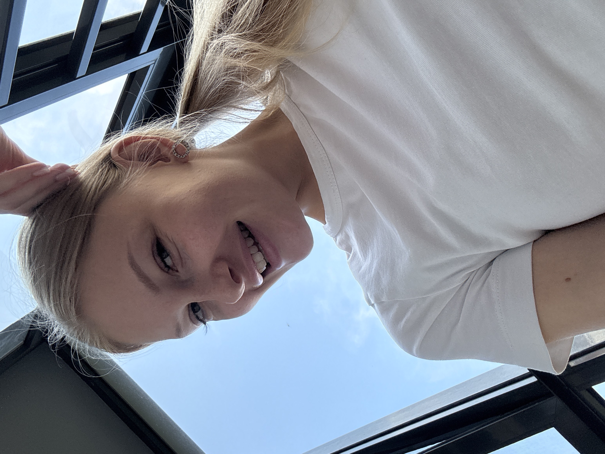 A young blonde woman with her hair in a ponytail smiles into the camera, her head tilted slightly upwards. She is wearing a simple white t-shirt. Her right hand is raised and touching her hair. She has a small silver hoop earring in her right ear. The background is a bright blue sky with some clouds, and dark, angular structures that suggest an urban environment, possibly a balcony or rooftop overlooking the city of Perm, Russia. The perspective is from below the woman, looking up at her and the sky.