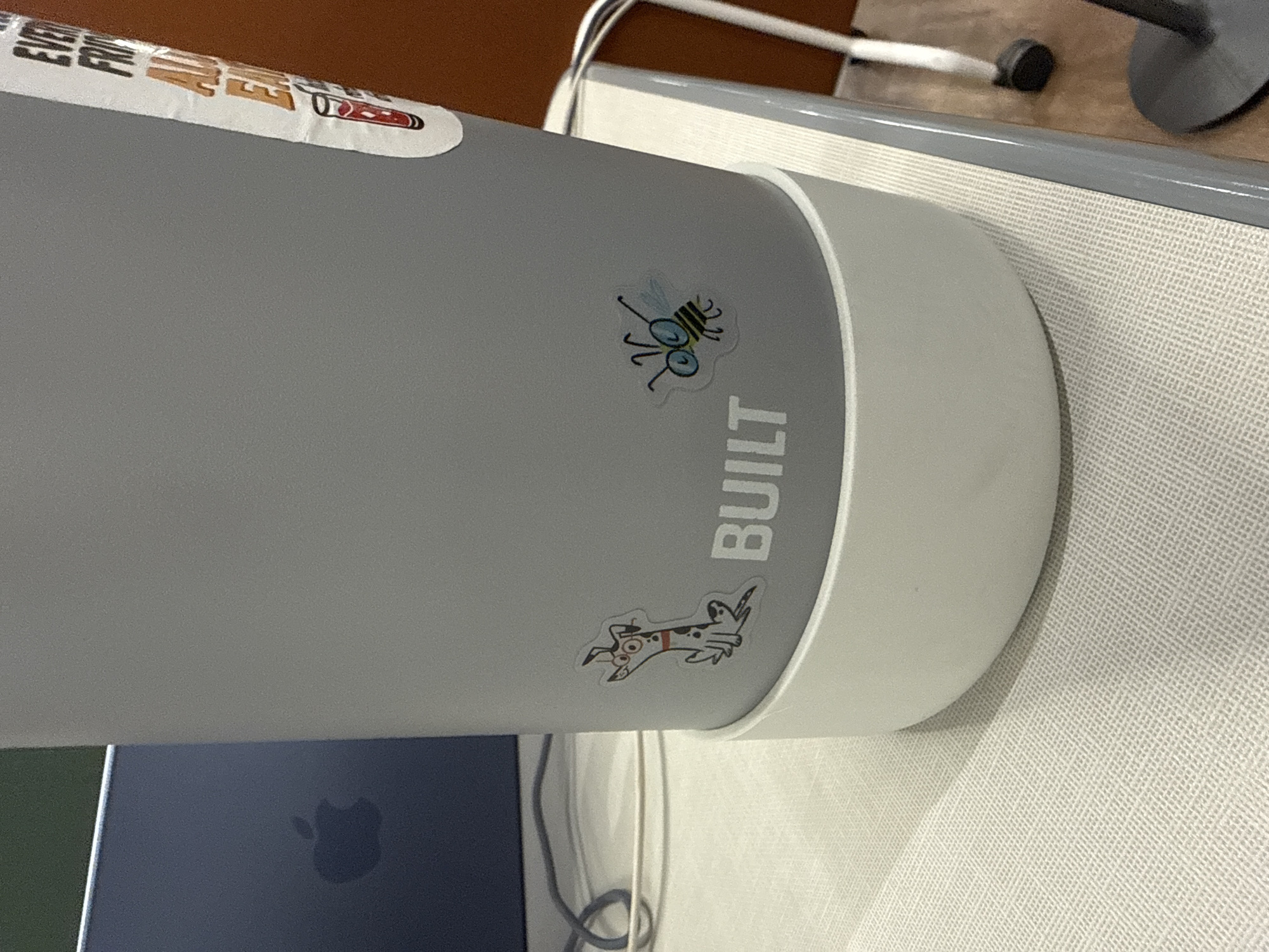 The image displays a close-up of a grey cylindrical object, likely a tumbler or water bottle, resting on a light-colored, textured desk surface in an indoor setting in San Andrés Cholula, Mexico. The object features a white base or rim.
Decorating the grey surface are two distinct stickers. One sticker depicts a cartoonish fly with large, round eyes and transparent wings. Below it, another sticker shows a whimsical, spotted dog, resembling a Dalmatian, in a running or jumping pose, wearing a red collar. Vertically aligned on the grey object, the brand name "BUILT" is visible, with the letters "BUI" clearly seen, followed by "LT" below.
Partially visible at the top edge of the grey object is a white label or sticker with some indistinct text and a small red and black graphic. In the lower left of the frame, the corner of a dark-colored laptop with a prominent Apple logo is visible. Various charging or data cables (white and black) are also present on the desk, near the laptop. The background includes a darker brown or orange surface in the upper left and a metal support structure, possibly from a chair or desk, in the upper right. No specific activity is taking place; the image is a still life of everyday items on a workspace.