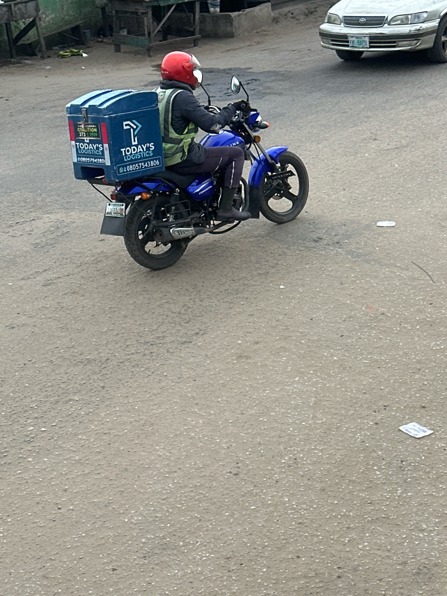 A male delivery person wearing a red helmet and a high-visibility yellow vest is riding a blue motorcycle on a paved road. The motorcycle has a large blue cargo box attached to the rear, displaying the "TODAY'S LOGISTICS" logo and a phone number. The delivery person appears to be moving forward. The background includes a silver car, some roadside structures, and debris on the ground, suggesting an urban or semi-urban environment, possibly in Ajara, Nigeria.