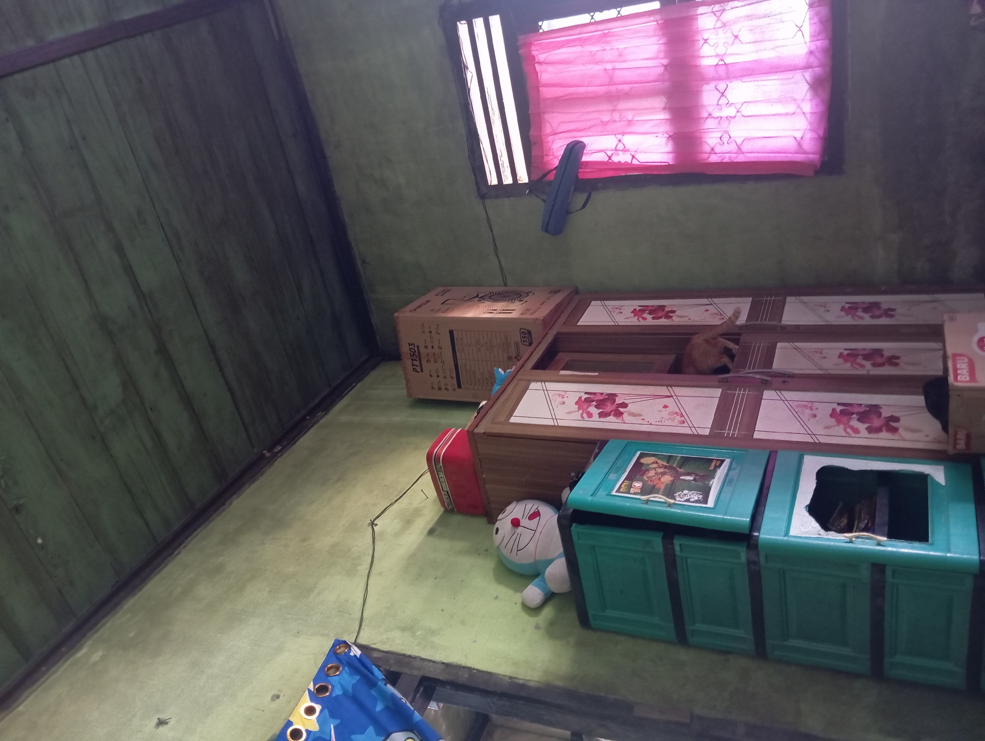 The image depicts a cluttered interior room, likely a bedroom or storage area, within a modest dwelling in Rejosari, Indonesia. The walls are painted a faded green, and the ceiling is constructed from wooden planks. Prominently featured are several storage units: a tall, green plastic multi-drawer cabinet with images of cartoon characters on one drawer, and a wooden wardrobe with floral patterns on its doors. On top of the green cabinet sits a plush toy resembling the character Doraemon. To the right of the wardrobe is a cardboard box labeled "PT1503". A pink curtain hangs over a window on the right side of the frame. A cat, appearing to be ginger-colored, is partially visible peeking out from an opening in the wooden wardrobe. On the left side of the image, a blue curtain with star patterns is draped, partially obscuring a doorway or opening. The overall impression is of a lived-in, possibly somewhat unkempt, domestic space.