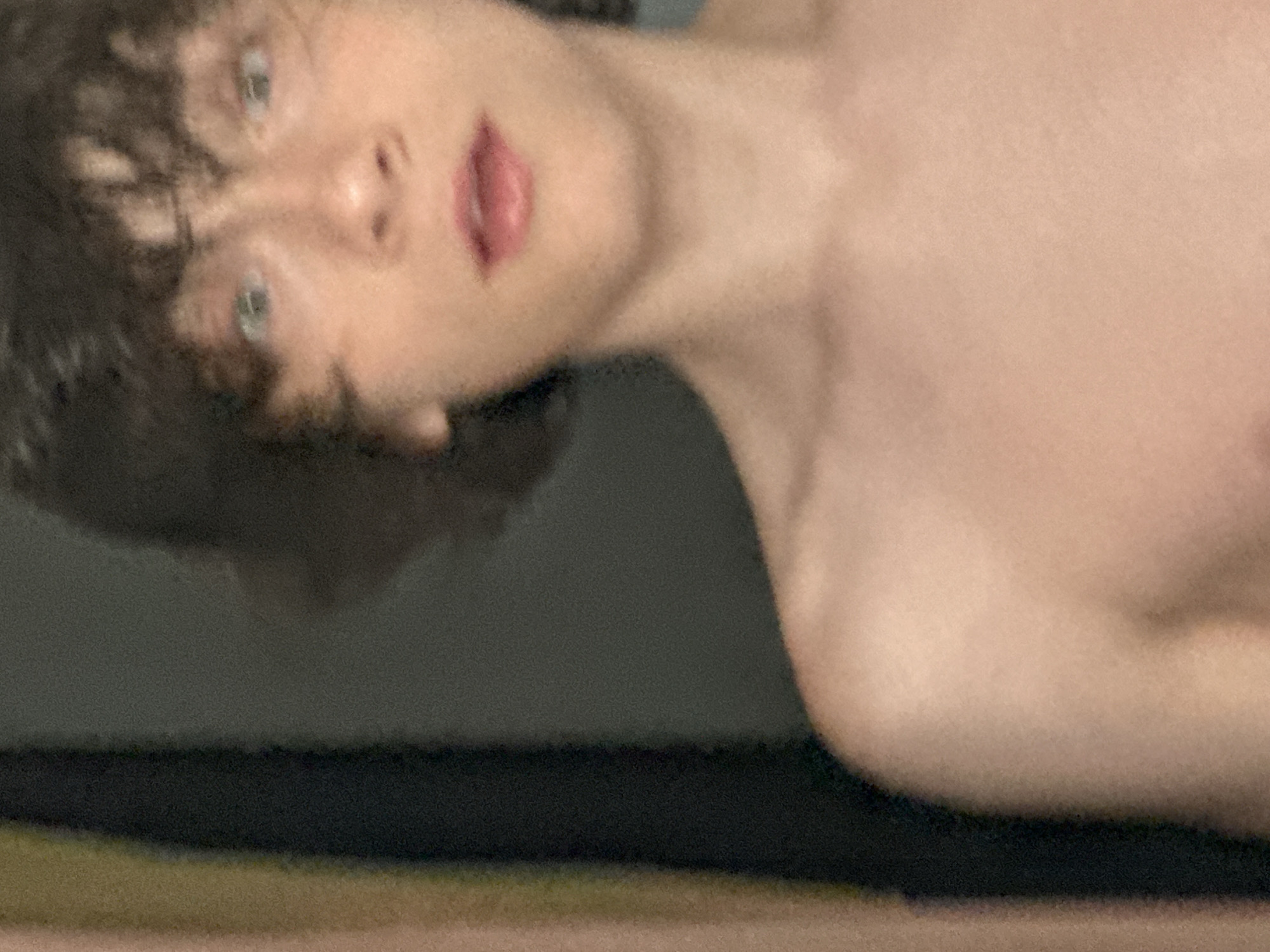 The image is a close-up, slightly blurred, waist-up shot of a young person with dark, curly hair. The person is looking slightly up and to the right, with their mouth open. Their skin appears fair, and their eyes are light-colored. The background is dark and out of focus, with vertical bands of lighter and darker colors visible on the left side of the frame. There is no discernible information in the image that would allow for the identification of a specific location in Sochi, Russia, or any other geographical area.