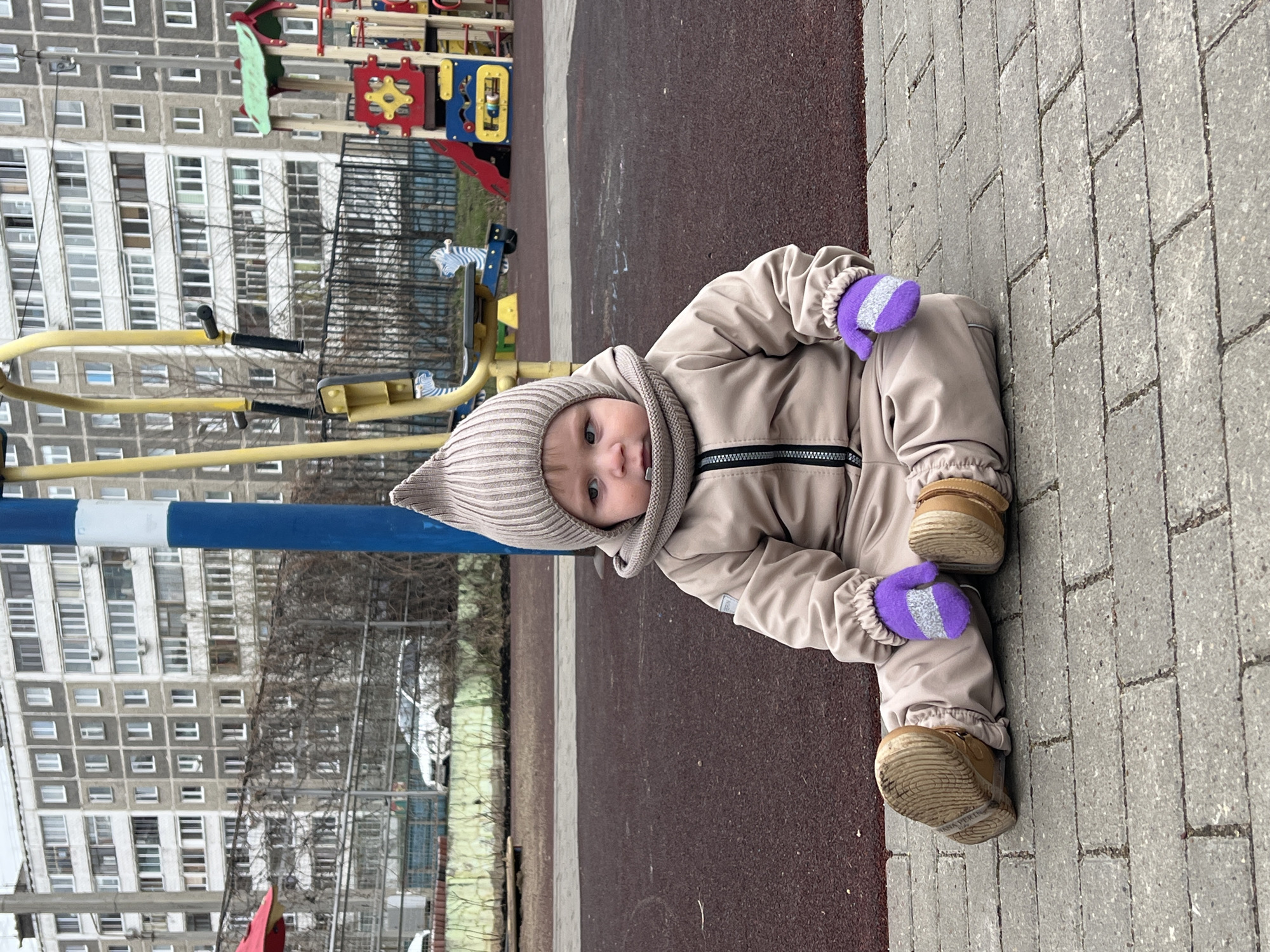 A young infant, dressed in a beige/tan full-body snowsuit, a matching hooded hat, purple mittens with a white stripe, and tan shoes, sits on the ground at a playground. The infant, with blue-grey eyes, looks directly at the camera with a neutral or slightly curious expression.
The child is positioned at the boundary between a dark reddish-brown, possibly rubberized, playground surface and a grey paved walkway made of rectangular blocks arranged in a herringbone pattern. In the background, yellow and blue playground equipment is partially visible, including what appears to be a swing set or climbing frame. Behind the playground, a tall, multi-story residential building, likely an apartment block with numerous windows, stands prominently. Its grey concrete facade is characteristic of architecture found in Perm, Russia. The overall lighting suggests it is daytime, and the infant's warm attire indicates cool or cold weather, possibly overcast. On the sole of the infant's right shoe, the brand name "HANNA PERKINS" is visible. The image is oriented with the scene rotated 90 degrees clockwise.