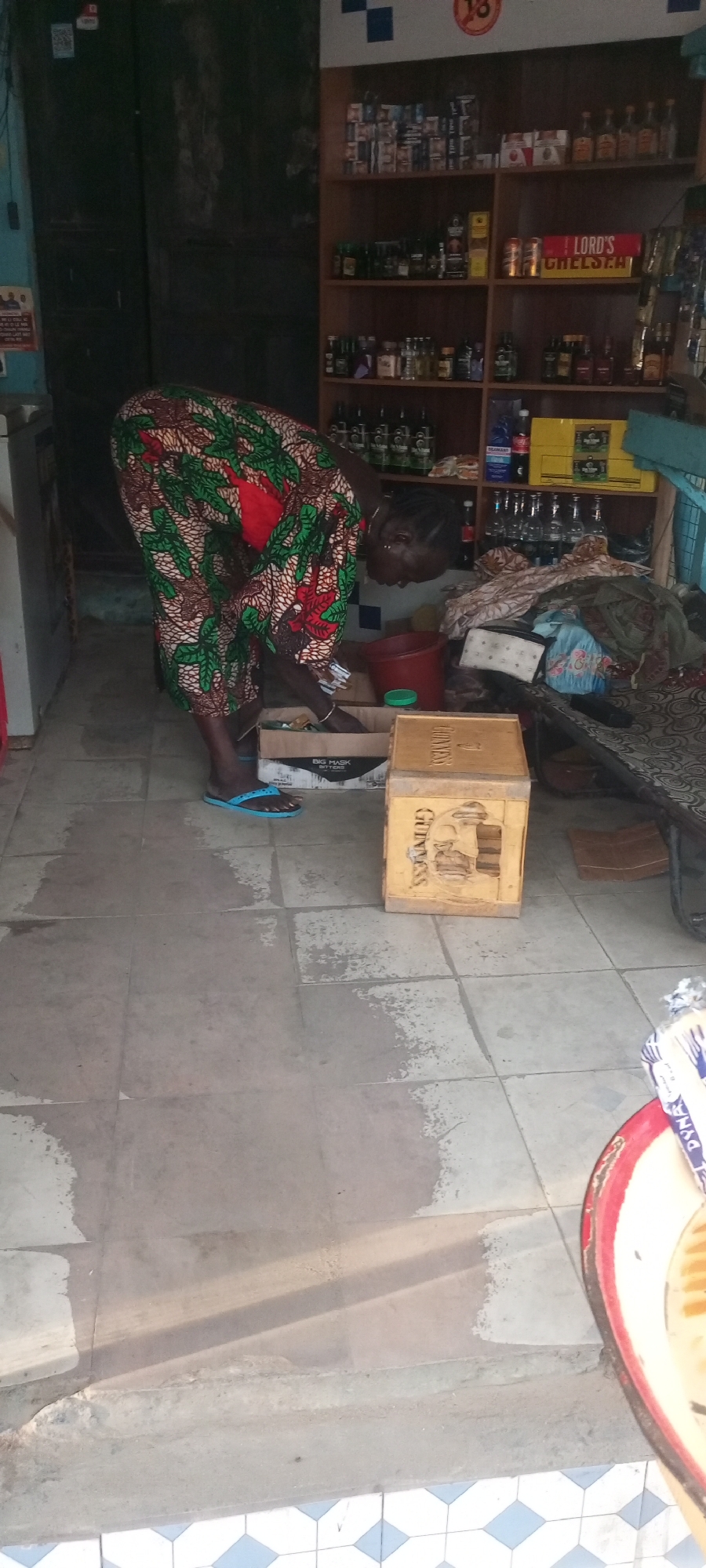 The image shows a woman in Adankolo, Nigeria, bending over a cardboard box labeled "BIG MASK BITTERS" inside a shop. She is wearing a patterned dress in brown, green, and red, and blue flip-flops. Next to the box is a wooden crate with "GUINNESS" printed on it. Behind the woman is a shelf stocked with bottles, boxes, and other items. A red bucket is visible on the shelf, and a bench covered with a patterned cloth is to the right. The floor is tiled in a light grey color with patches of discoloration, and a blue-and-white tile pattern is at the bottom of the frame. Sunlight is streaming into the shop.