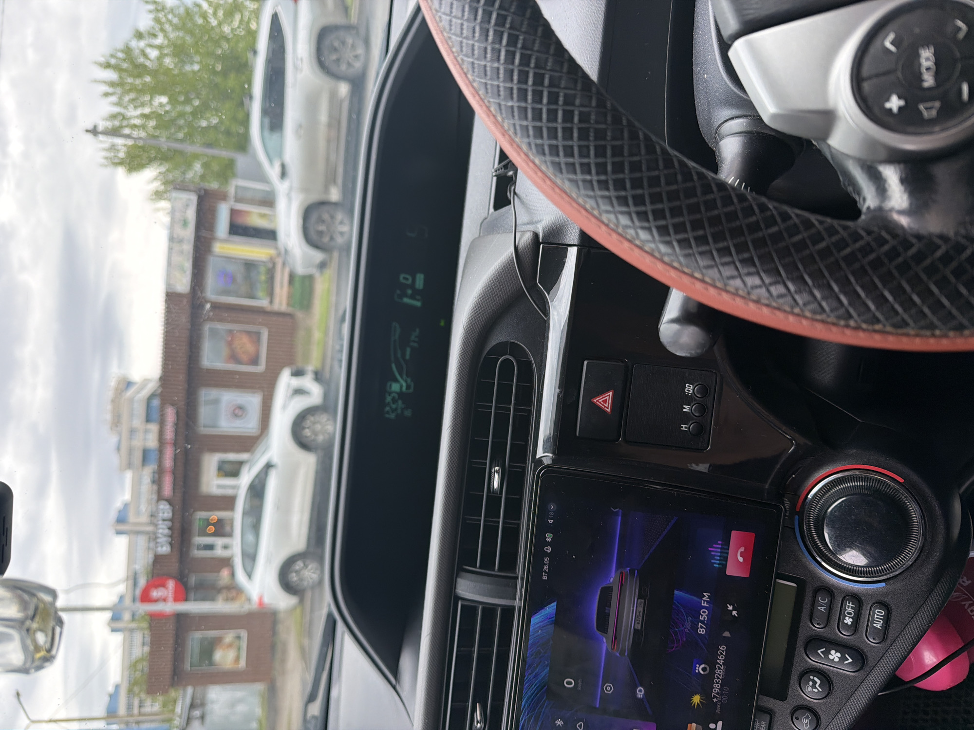 The image shows the interior of a car, specifically the dashboard, steering wheel, and center console. The car appears to be parked or stopped, as seen through the windshield, where a building with the sign "БУХТЕР" (BUKHTER) is visible. "Бухтер" is a store of alcoholic beverages. The building is located in Krasnoyarsk, Russia. The radio is tuned to 87.50 FM. The time is 26:05.