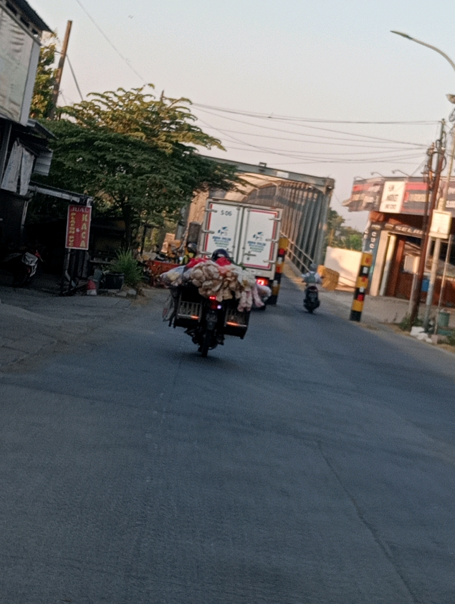 The image depicts a street scene in Soko, Indonesia, with a focus on a motorcycle laden with goods. The motorcycle, seen from behind, is carrying a large, bulging sack on its rear rack, secured with straps. A person is riding the motorcycle, wearing a red shirt and a helmet. The motorcycle is on an asphalt road, with a slight incline. To the left, a building with a corrugated roof and a sign that reads "JUAL KACA" (meaning "GLASS FOR SALE") is visible. Several trees are present in the background, with lush green foliage. Further down the road, a white truck with the number "506" on its side is parked, and another motorcycle is also visible. The sky is clear, suggesting a daytime setting. The overall environment appears to be a somewhat commercial or residential area in a city or town.