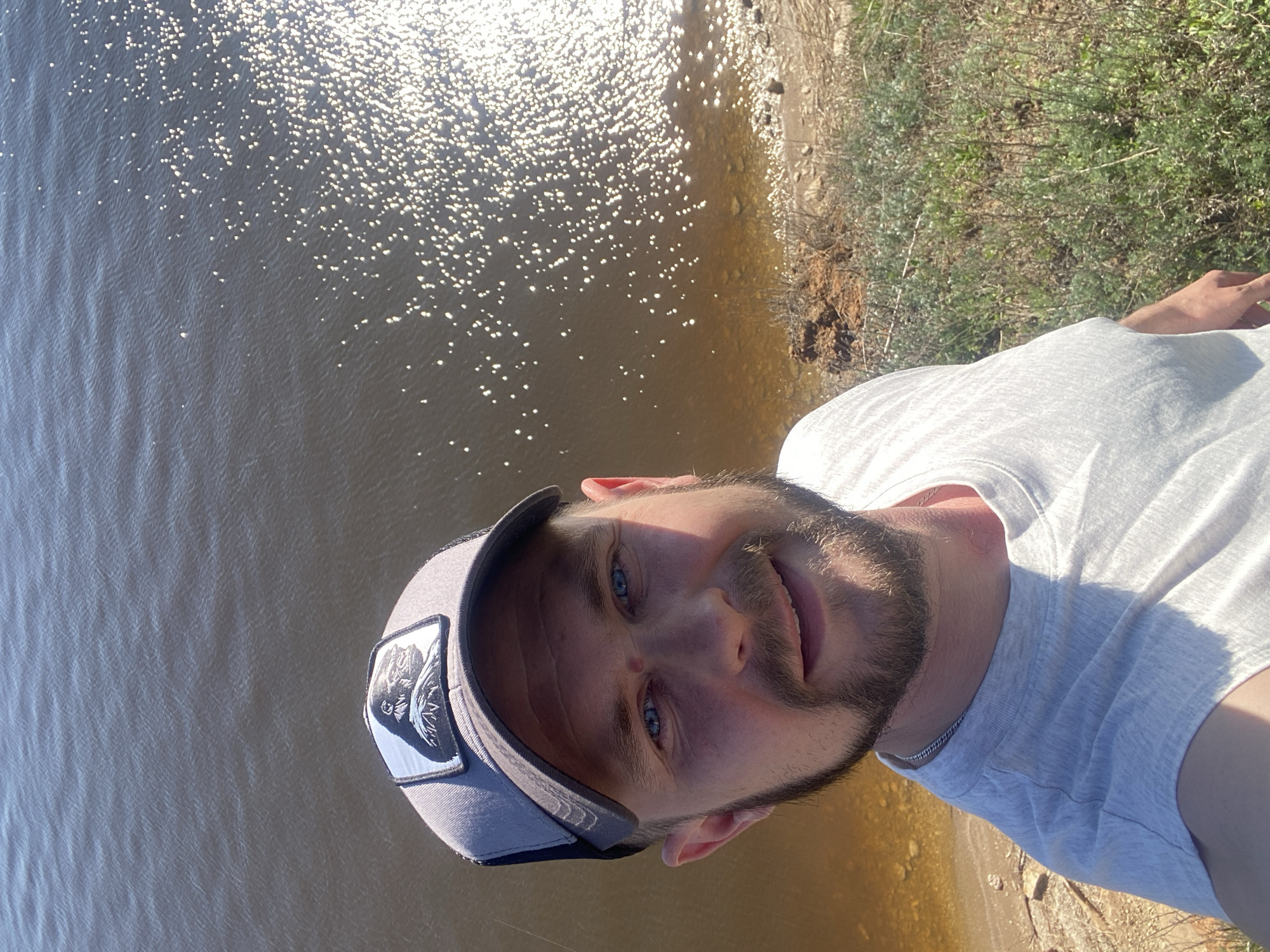 The image shows a selfie taken outdoors, likely by a man with fair skin, blue eyes, and a beard, wearing a white t-shirt and a light gray baseball cap with a patch on the side. The background includes a body of water with reflective sunlight, and some vegetation along the shoreline. The photograph appears to have been taken in daylight with bright lighting conditions. Based on the details given, the location is Шуран, Russia.
