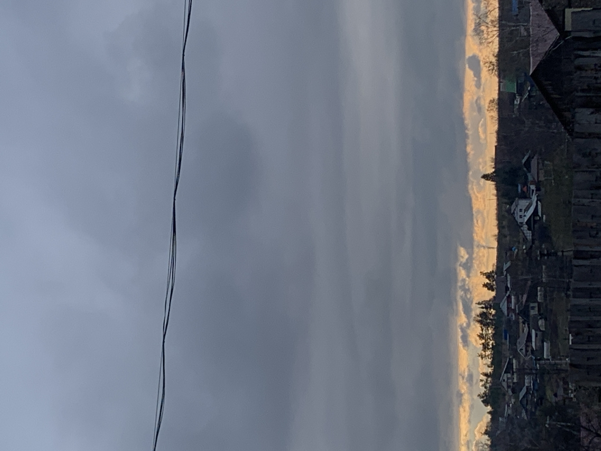 The image displays a landscape scene, rotated such that the horizon runs vertically, with the sky on the left and the land on the right. When viewed correctly (rotated 90 degrees counter-clockwise), the sky occupies the upper two-thirds of the frame, dominated by heavy, uniform gray clouds, with some lighter patches.
Along the horizon, a distinct horizontal band of golden-orange light cuts through the cloudy sky, hinting at either sunrise or sunset. Below this band of light, the silhouette of a village or small town spreads across the lower third of the image. Various buildings with dark roofs are visible, interspersed with bare trees and sparse vegetation, creating a somewhat rustic or rural impression.
In the upper left corner, two dark, thin lines, likely power or telephone wires, stretch horizontally across the cloudy expanse. There are no visible people or discernible activities taking place. The overall impression is one of quietude, with the dramatic contrast between the somber clouds and the intense glow of the low sun setting a distinct mood. No text is visible within the image. The location is Фоки, Russia.