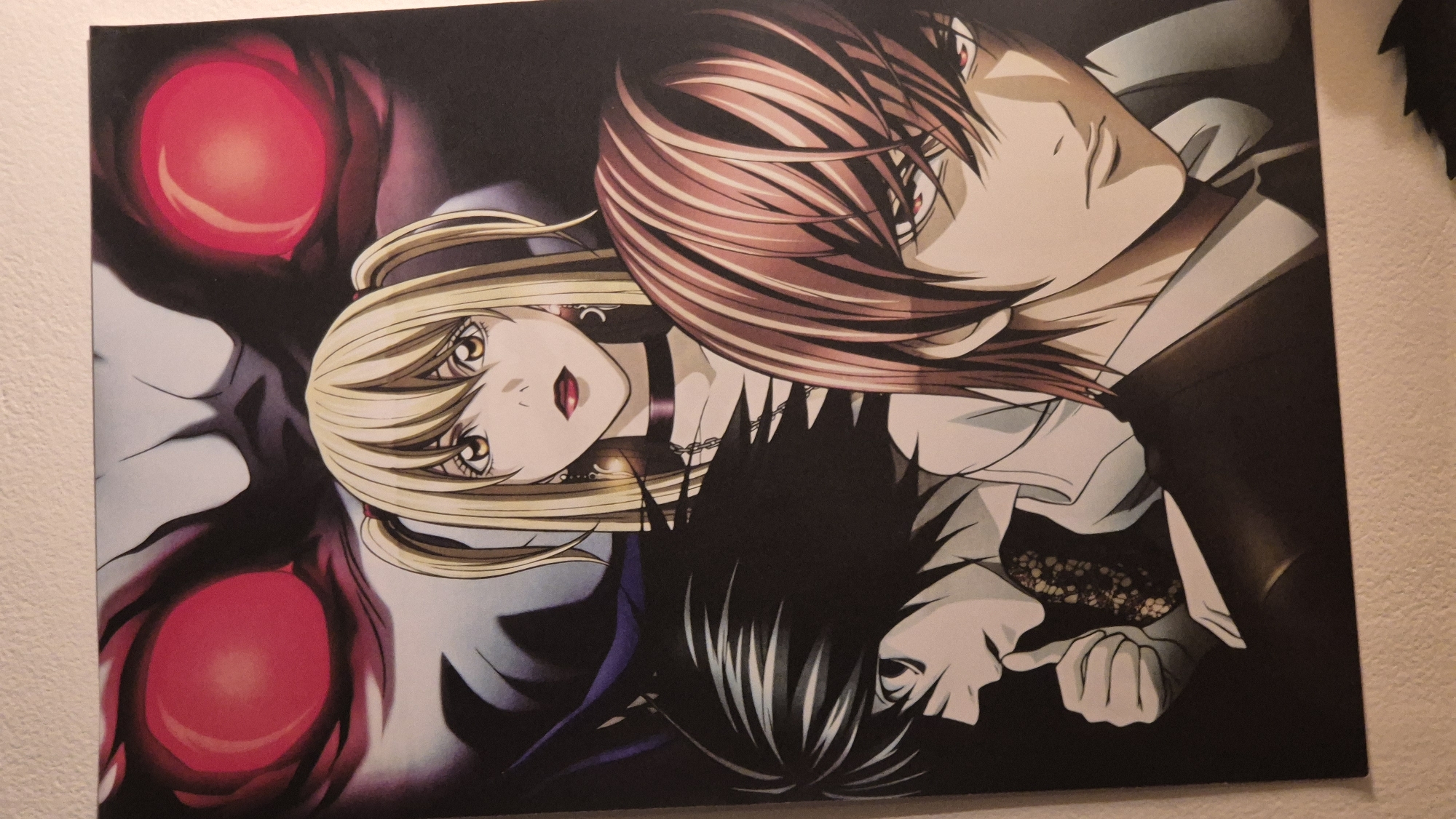 This is a close-up, eye-level, indoor shot of a poster featuring characters from the anime series Death Note. The poster is vertically oriented. Dominating the top portion of the poster is a large, shadowy figure with prominent, glowing red eyes. Below this figure, from left to right, are three human characters. On the left, a male character with spiky black hair and dark eyes is depicted in profile, looking to the left and with his index finger touching his lips. In the center, a blonde female character with pigtails, large golden eyes, red lipstick, and wearing a choker is looking directly at the viewer. To the right of her, a male character with reddish-brown hair and red eyes is shown from the chest up, looking towards the viewer's left. The poster is mounted on a textured, light-colored wall. The lower portion of the poster is not fully visible, but the overall impression is a dramatic and intense composition of the characters. The image does not depict any elements that would allow for identification of a specific location within the city of Sofia, Bulgaria.