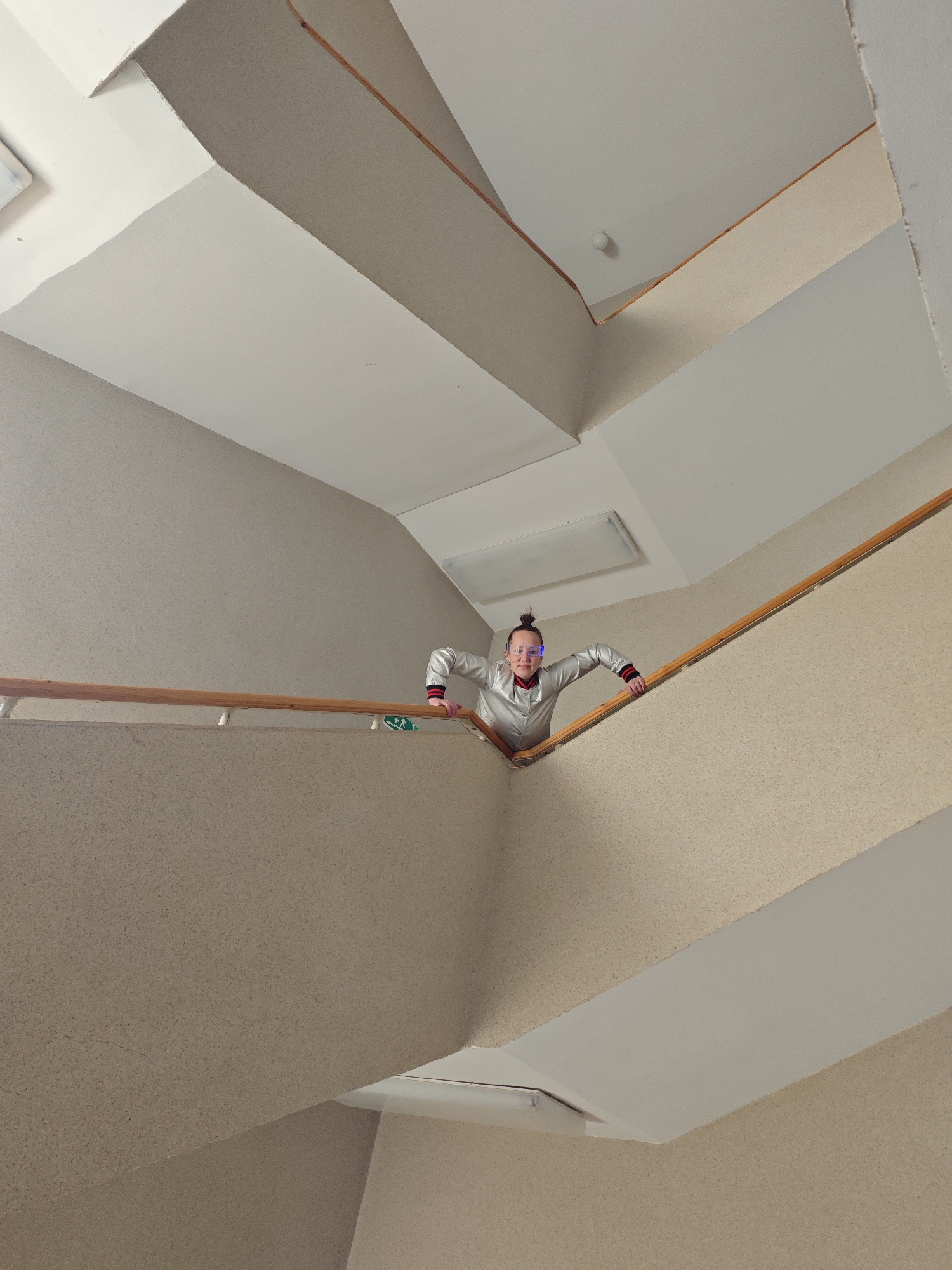 A low-angle, long shot captures a person in a building's stairwell. The stairwell is constructed of beige-colored walls and ceilings with white trim along the ceiling edges. A wooden handrail runs along the wall. A person with their hair in a bun is peering over the edge of the stairwell, holding onto the handrail. The person wears a silver-colored jacket with red and black trim at the cuffs and is also wearing glasses with blue lights. The lighting is from above, illuminating the stairwell. The location is identified as Марковский, Russia.