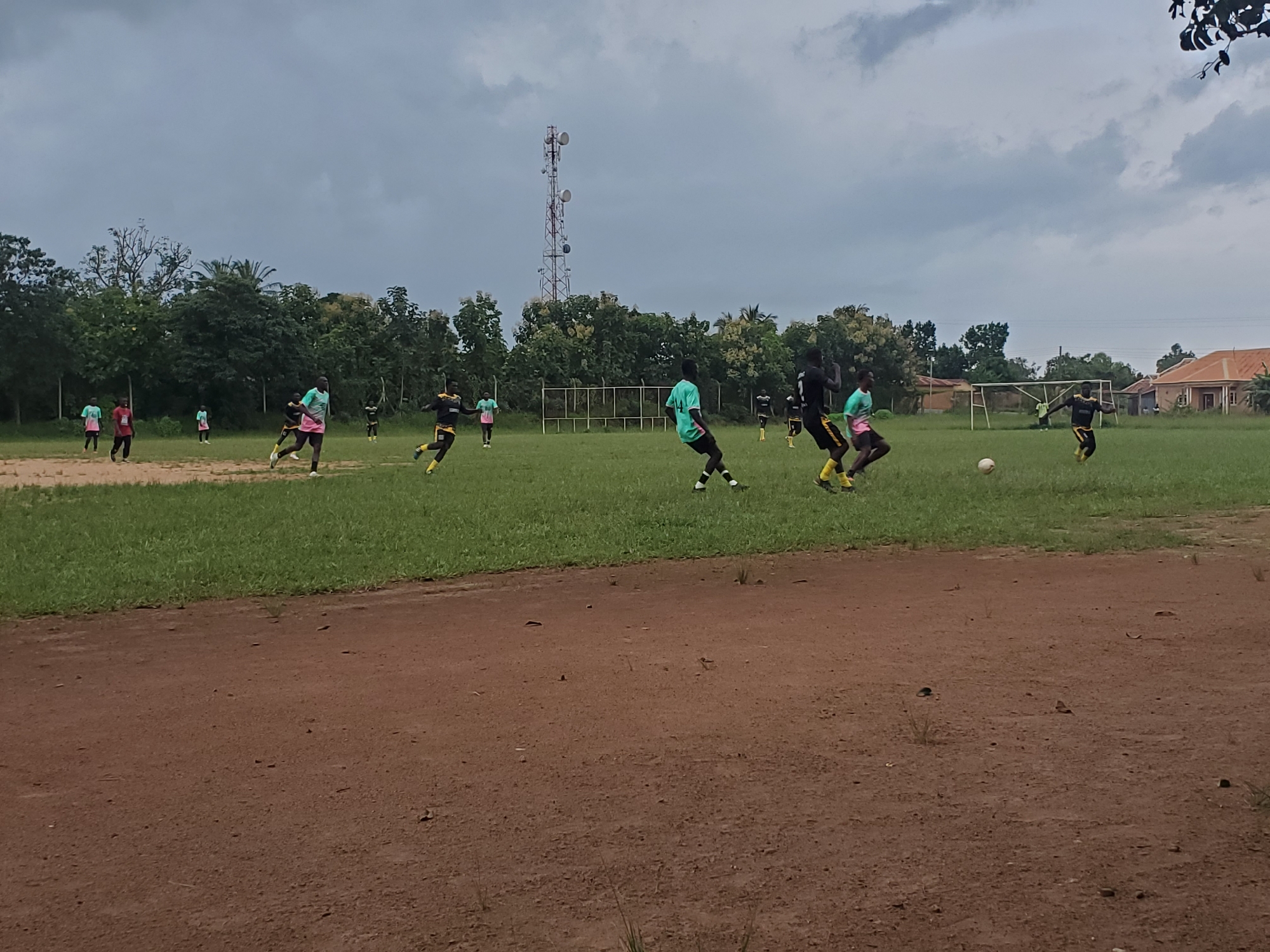 The image depicts a soccer game in progress on a grassy field in Miango, Nigeria. Several young men, dressed in soccer uniforms, are actively engaged in the game. Some are in motion, kicking or preparing to kick a soccer ball, while others are running or positioned on the field. The field is bordered by trees and some buildings are visible in the background, along with a cell phone tower. The sky is overcast with clouds. The foreground of the image shows a dirt track.
