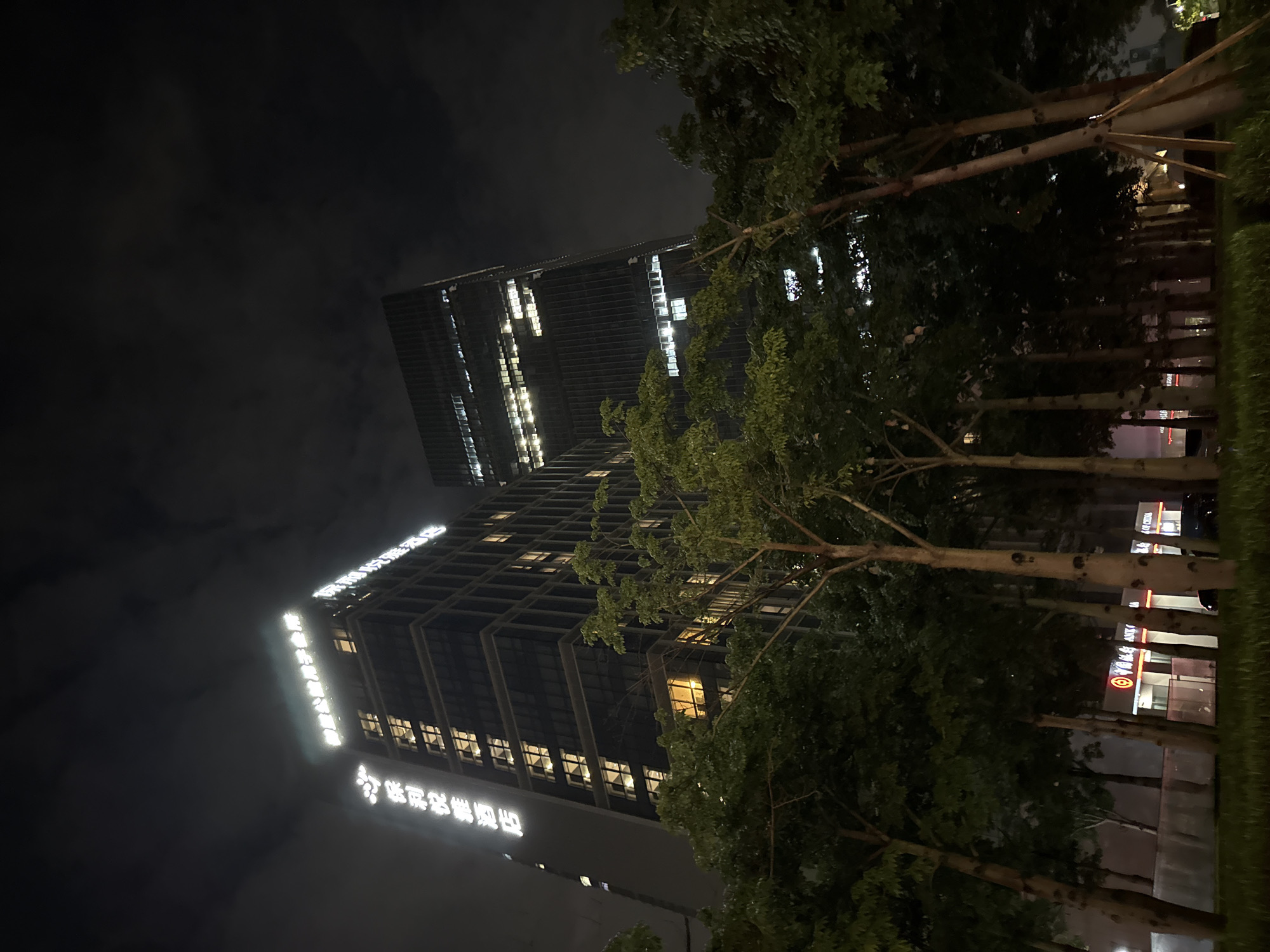 A nighttime view of a tall, modern skyscraper in Haizhu District, China. The building is illuminated by numerous lit windows, casting a warm glow against the dark sky. The facade of the building features prominent white lettering, which is partially obscured by trees and the angle of the shot. Some of the visible text appears to be in Chinese characters.
In the foreground, a line of trees with sparse green foliage stands between the viewer and the building. Their branches extend upwards, partially obscuring the upper sections of the skyscraper. The ground around the trees is covered in grass and some paved areas. Beyond the trees, at the base of the building, there are more illuminated signs, some of which appear to be commercial or corporate logos. One visible logo is a red circle with a white design inside. The overall scene suggests an urban environment at night, with the building representing a significant architectural landmark. The presence of trees indicates a landscaped area, perhaps a park or public space adjacent to the building. The sky is dark and cloudy, with some atmospheric haze contributing to the moody nighttime ambiance. There are no people visible in the frame.