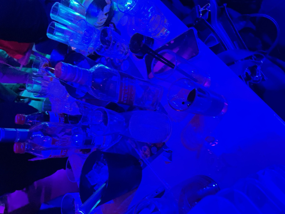 The image presents an indoor scene bathed predominantly in a deep, electric blue light, characteristic of a bar, nightclub, or lounge setting. The primary subjects are numerous glass bottles and various barware items arranged on a light-colored surface, likely a bar counter or shelving.
Several bottles of varying shapes and sizes are visible, many appearing empty. A prominent bottle in the mid-left area, lying horizontally, is identifiable as a "JOHNNIE WALKER RED LABEL" whisky bottle, with its distinct text clearly readable despite the blue illumination. Other bottles have red caps, and one smaller bottle lower-left contains text that appears to be in an Asian script, though it is too blurry to decipher. Interspersed among the bottles are clear drinking glasses, some inverted, and metal bar tools such as a black bar spoon and a metallic jigger or measuring cup, indicating a functional bar setup.
The scene suggests a vibrant, artificial ambiance, with the intense blue lighting creating reflections and highlights on the glassware. Hints of other colored lights, such as green and purple, can be seen in the background, adding to the dynamic atmosphere. The time of day is clearly night, as indicated by the reliance on artificial lighting. No specific activity is depicted, but the arrangement of bottles and bar tools implies either active drink service or a display within a venue dedicated to alcoholic beverages. While no individuals are distinctly visible, blurred shapes in the background might suggest human presence. The overall impression is that of a modern and lively establishment in Drapetsona, Greece.