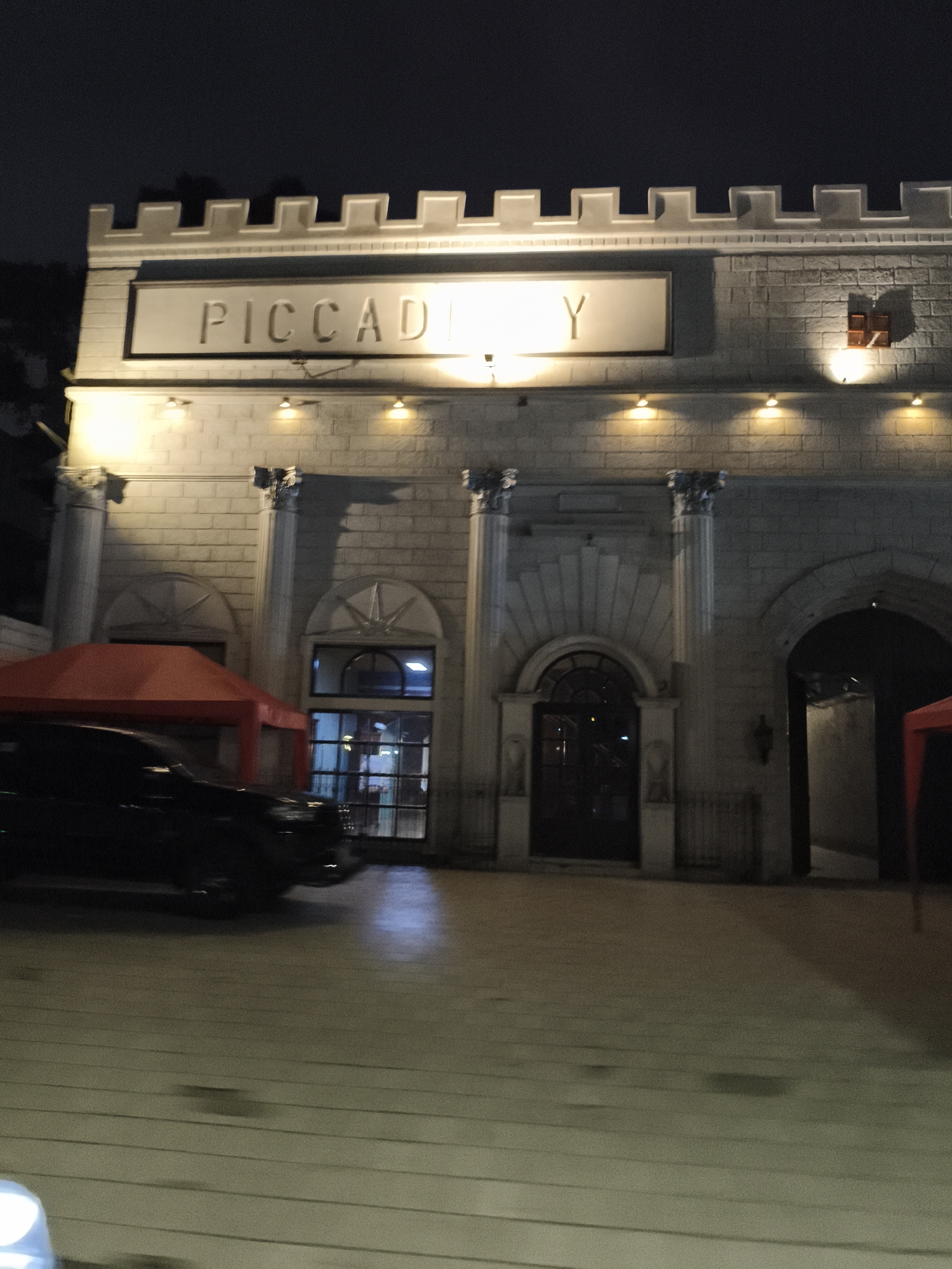 The image depicts the exterior of a building at night, illuminated by artificial lights. The building's facade is constructed of light-colored bricks and features architectural elements reminiscent of a castle, including a crenellated parapet at the top. A prominent sign above the entrance reads "PICCADILLY" in raised lettering. The building has several arched doorways and windows, with some of them displaying interior lighting. A red tent structure is visible on the left side of the building, and a dark-colored vehicle is parked in front of it. The ground in front of the building appears to be paved. The sky is dark, indicating it is nighttime. The overall setting suggests a commercial establishment, possibly a restaurant or entertainment venue. This location is identified as Piccadily, located in South Jakarta, Indonesia.