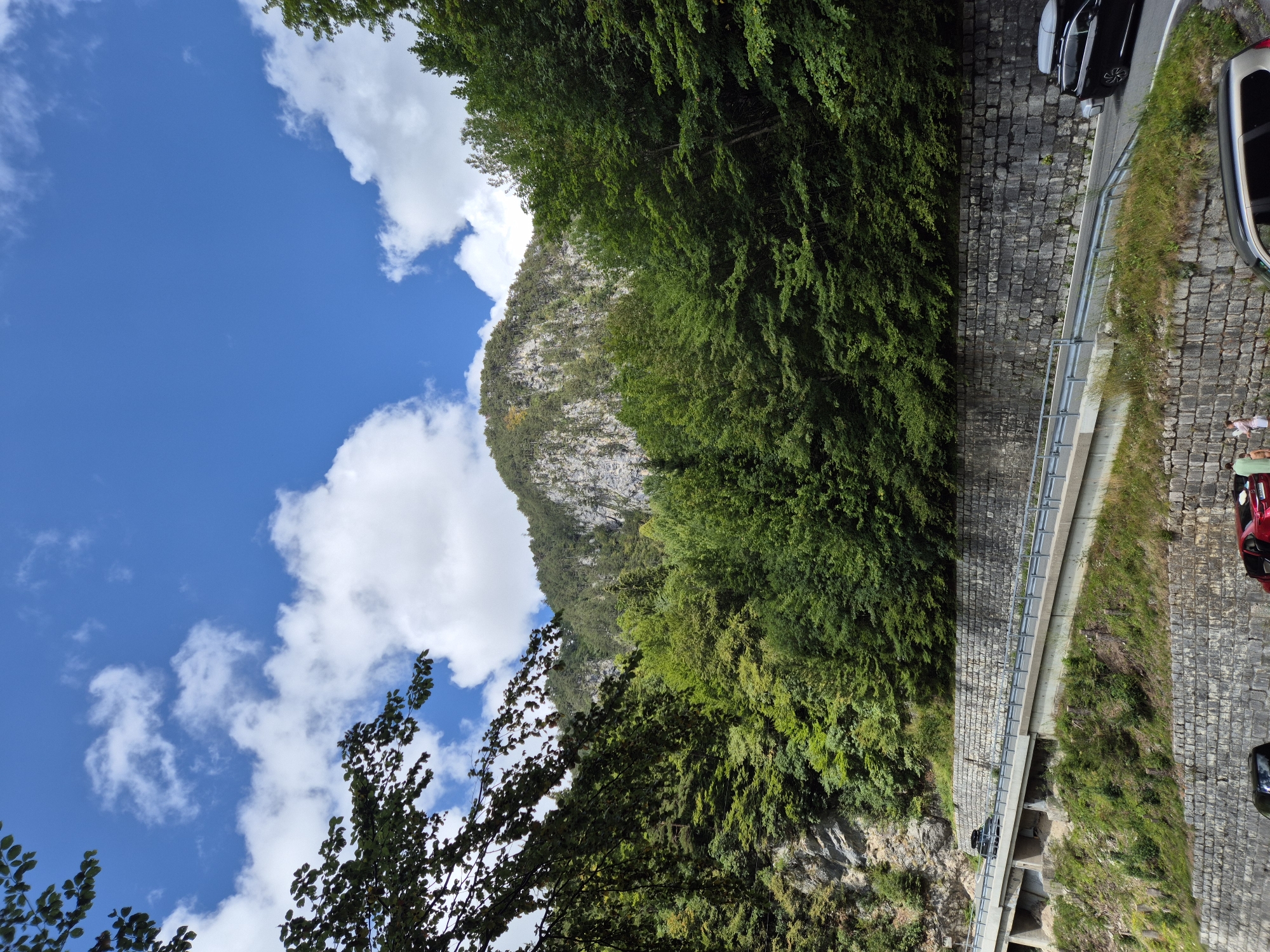 A paved road curves uphill, bordered by a stone retaining wall on one side and a metal guardrail on the other. A dark SUV is parked on the roadside to the right, with a roof box. Further up the road, another dark SUV is driving away from the camera. The road is surrounded by dense green trees and a rocky mountainside in the background. The sky above is blue with scattered white clouds. The scene appears to be a scenic route in a mountainous area, possibly near Ferlach, Austria, given the context.