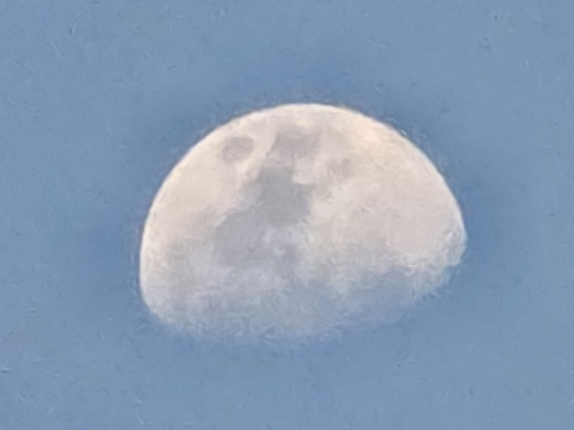 The image is a close-up, slightly blurry photograph of a crescent moon against a clear blue sky. The moon is illuminated on its right side, with the left side in shadow. The surface of the moon shows some visible craters and maria, though they are not sharply defined due to the image quality. The sky is a uniform light blue, with no clouds or other celestial bodies visible. There is a subtle grainy texture across the entire image, likely due to the camera's resolution or processing. The photograph does not contain any elements that would allow for the identification of a specific location in Yambol, Bulgaria, or any people, objects, actions, or interactions.