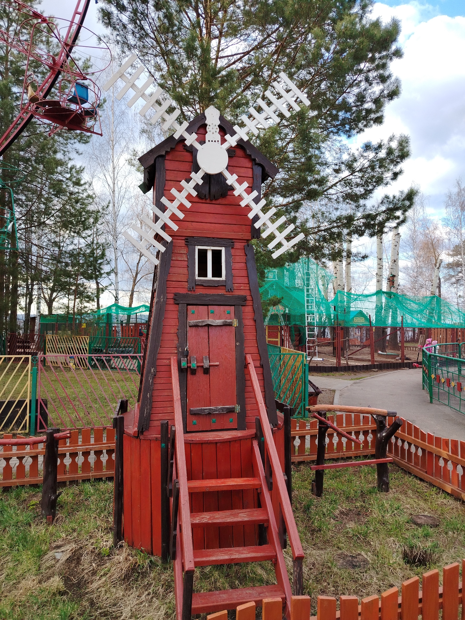 This image captures a vibrant outdoor scene at what appears to be a children's or amusement park in Chaikovsky, Russia. The central subject is a tall, red wooden decorative windmill with white blades, featuring a small window and a matching red door, surrounded by a low wooden fence. Red wooden stairs lead up to the windmill's door.
In the background, a variety of trees are visible, including green pine trees and slender white birch trees, some still bare. To the left, part of a red Ferris wheel-like structure with small cages is visible, suggesting an amusement ride. In the mid-ground, a large area is covered with green safety netting, indicative of a high ropes course or similar adventure playground, with a ladder visible inside. A paved pathway runs along the right side of the frame, bordered by colorful wooden fences in shades of red, orange, green, and yellow, designed in various patterns.
The scene is illuminated by natural daylight, suggesting it is daytime under a partly cloudy sky with patches of blue. No people are visible, but the presence of recreational equipment implies a setting for outdoor play and leisure. The grass around the structures shows patches of dry, brown vegetation amidst green, possibly indicating early spring or late autumn. No legible text is discernible within the image.