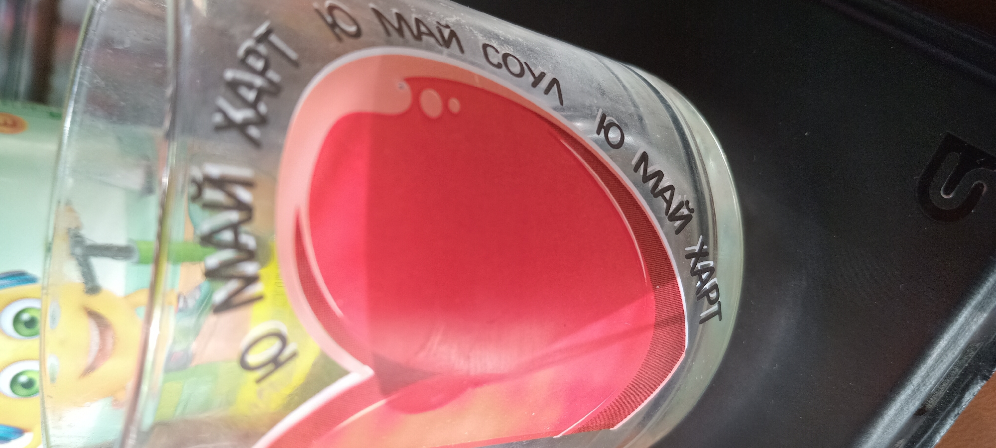 The image shows a clear glass with text and a heart design. The text "10 МАЙ ХАРТ" (10 May Hart) is printed on the glass in white lettering. Part of a yellow cartoon character with green eyes is visible in the background. The glass is resting on a dark, textured surface. The location in the city of Koviaky, Ukraine is not ascertainable from the image.
