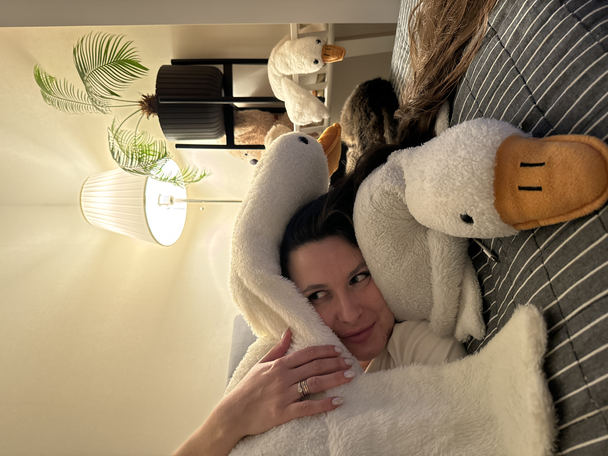 The image captures a woman relaxing indoors under warm, artificial lighting, likely in a bedroom or living room during the evening or nighttime. She is lying on what appears to be a bed or couch covered with a dark grey and white striped comforter. The woman has dark hair and a calm, content expression, looking towards the right side of the frame with a slight smile.
She is partially enveloped by a large, white, fluffy object, which could be a blanket, a hooded garment, or a substantial plush toy, designed to resemble a duck or goose. Its head, with an orange beak and embroidered eyes, is visible near her right shoulder. Her left hand, adorned with multiple rings, rests on this soft, white material.
Several other white duck or goose plush toys are scattered in the background; one is visible near her head on the striped blanket, and at least two smaller ones are perched on a dark shelf behind her. A portion of a dark-furred animal, possibly a cat, is partially visible behind her head, obscured by her hair and the blanket.
On the left side of the frame, behind the woman, a faux or real potted plant with green fronds, resembling a palm, sits in a black, ribbed planter on the same shelf. Below the plant, a white pleated lampshade emits a soft, inviting glow. A white charging cable is also discernible on the striped blanket. There is no visible text in the image. While the location context is given as Akulovo (Gp Kubinka), Russia, no specific visual cues in the image confirm this.