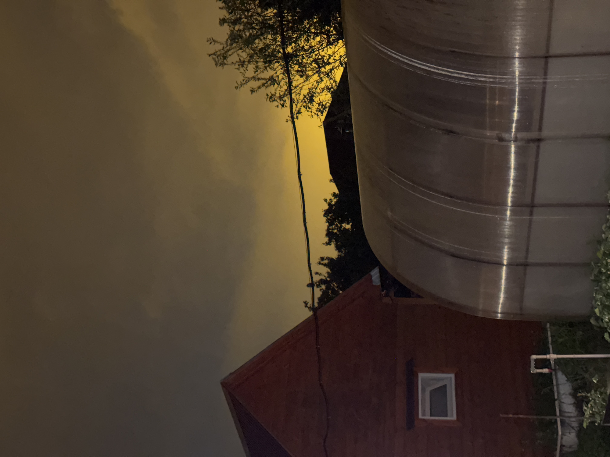 The image depicts an exterior nighttime scene in what appears to be the city of Mayak, Russia, given the context. The focus is on a red brick building with a single, small square window with the dark interior space visible through it. A large, shiny, cylindrical structure, possibly a metal storage tank or silo, dominates the right side of the frame. Overhead electrical wiring is visible against the background sky and near tree branches. The sky appears to be overcast and illuminated by an amber glow, suggesting artificial lighting. The ground is obscured by foliage.