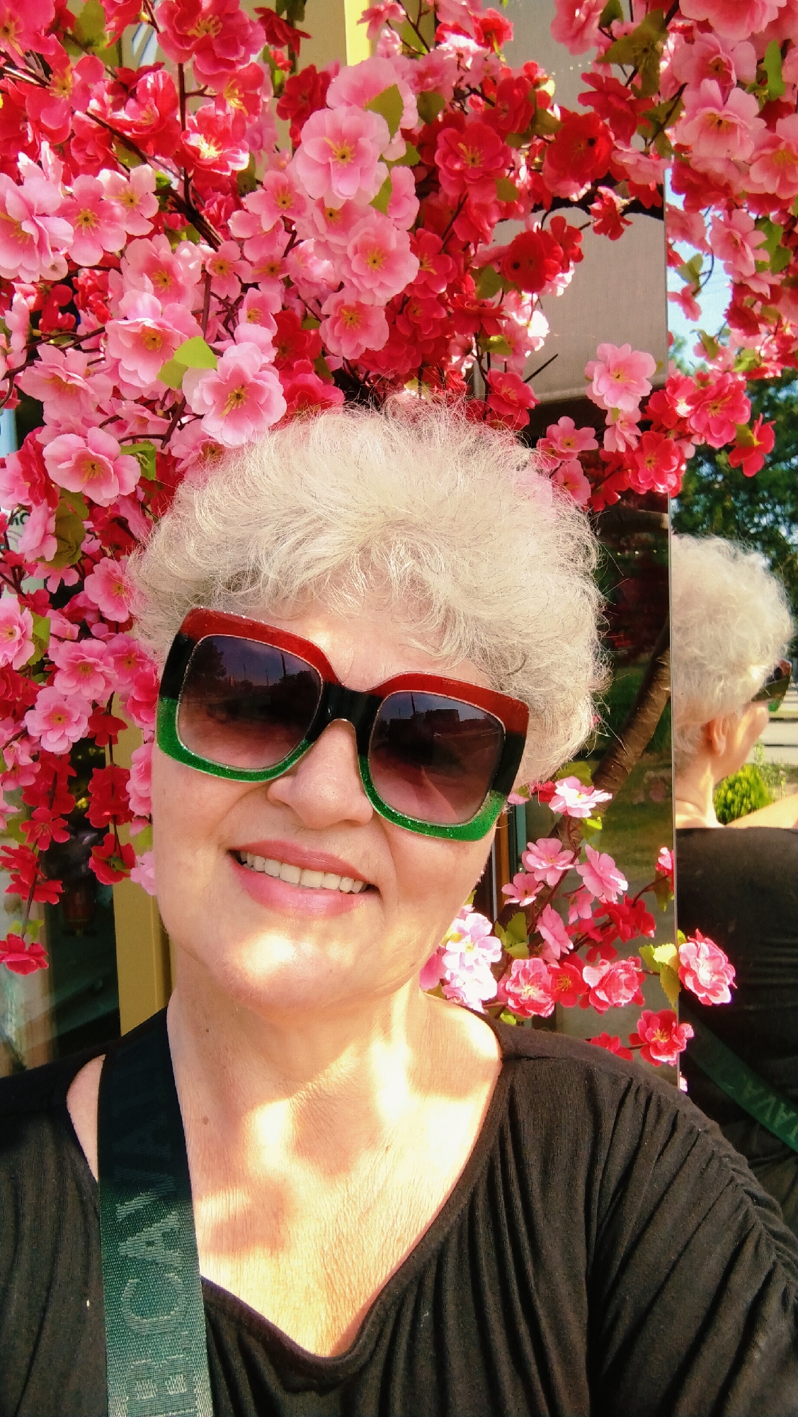 A selfie features an older woman with short, curly white hair and large, colorful sunglasses posing in front of a wall adorned with artificial pink and red cherry blossoms. The woman is wearing a black shirt with ruffled sleeves. In the reflection of a glass pane behind her, her back is visible, and she is wearing sunglasses and a black shirt. The setting appears to be outdoors, with hints of greenery and a building visible in the background reflection. The woman is smiling, and the overall impression is cheerful and vibrant due to the colorful blossoms. The provided context indicates this scene is located in the city of Horishni Plavni, Ukraine.