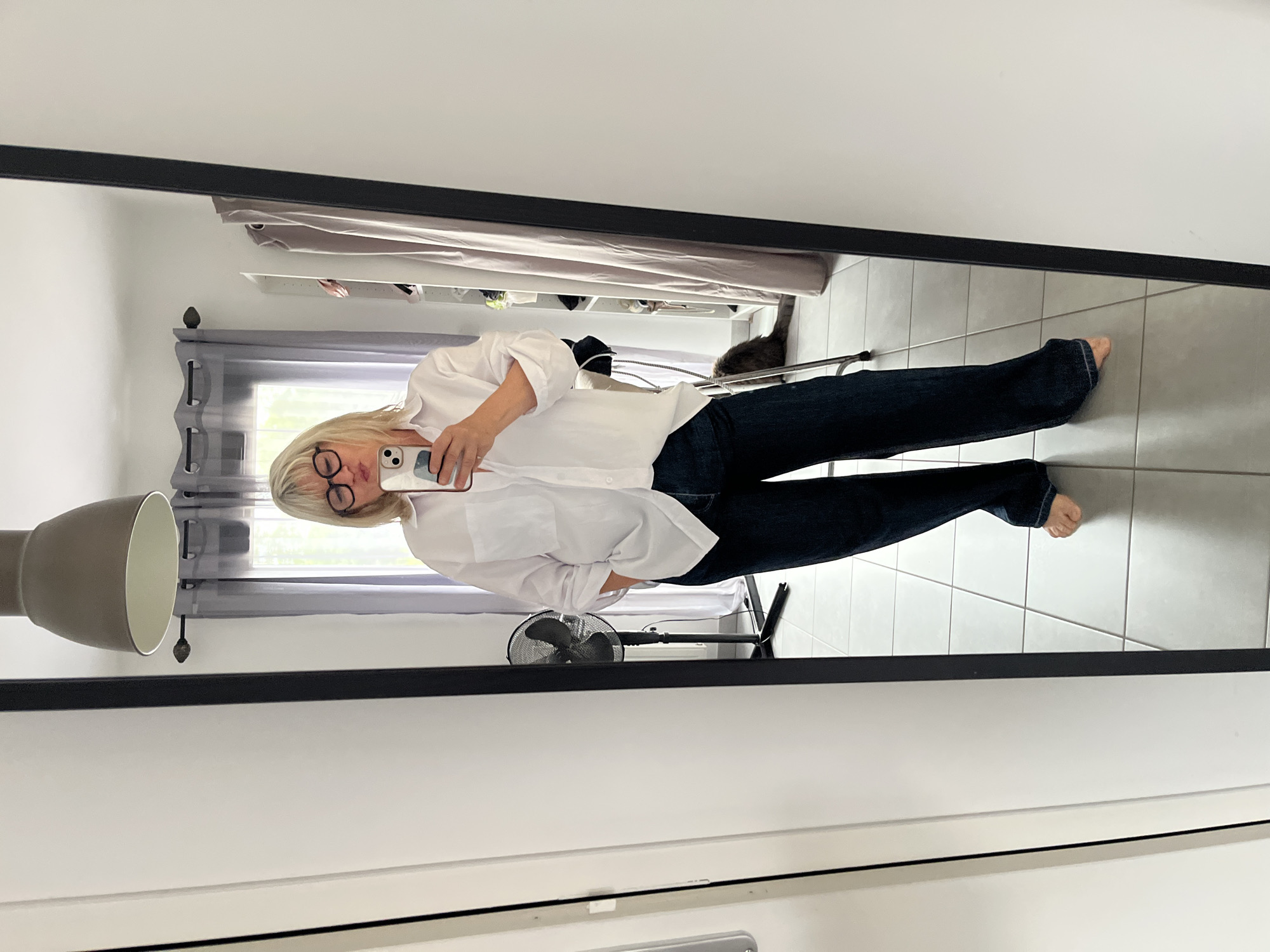 The image depicts a woman with blonde hair and glasses, dressed in a white shirt and jeans. She is holding a smartphone, likely taking a picture of herself in a mirror. The reflection reveals a room with light-colored tile flooring. Visible within the mirror's reflection are a floor lamp, a fan, a window with grey curtains, a clothing rack with various items, and a grey cat. The image was taken indoors in Chambœuf, France.