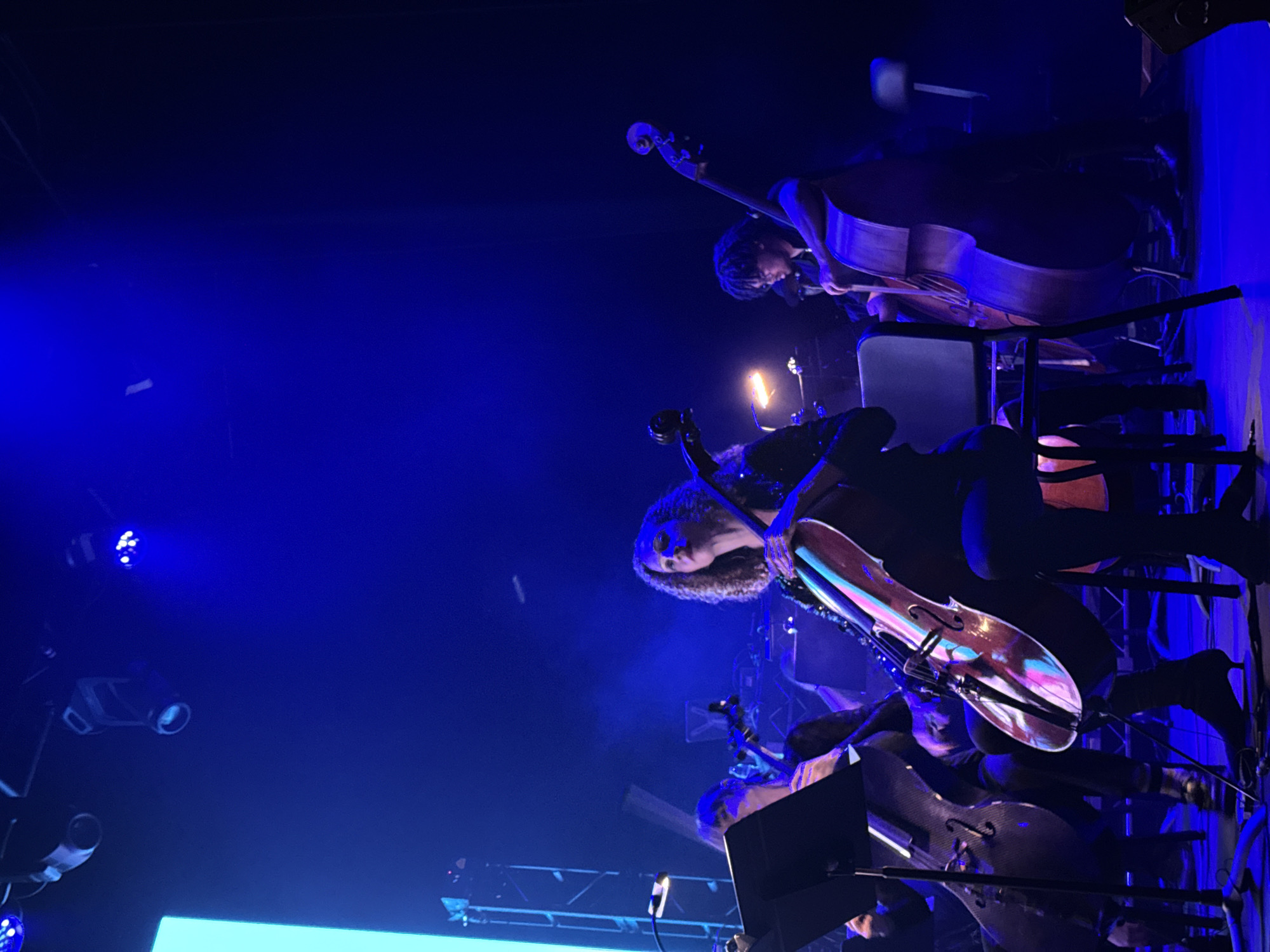 In Los Angeles, California, USA, an orchestra performs on a stage illuminated by vibrant blue lighting. Three cellists are visible, positioned from left to right. The cellist on the far left is mostly obscured, with only her left hand and a portion of her cello visible. The central cellist, a woman with curly hair, plays her cello, which is reflecting the colorful stage lights. To the right, a man with dark, curly hair and glasses plays a double bass. The stage is equipped with professional lighting fixtures and appears to be set up for a performance, with music stands and chairs for the musicians. The overall ambiance is that of a live concert, characterized by the dramatic lighting and the focused engagement of the musicians with their instruments.