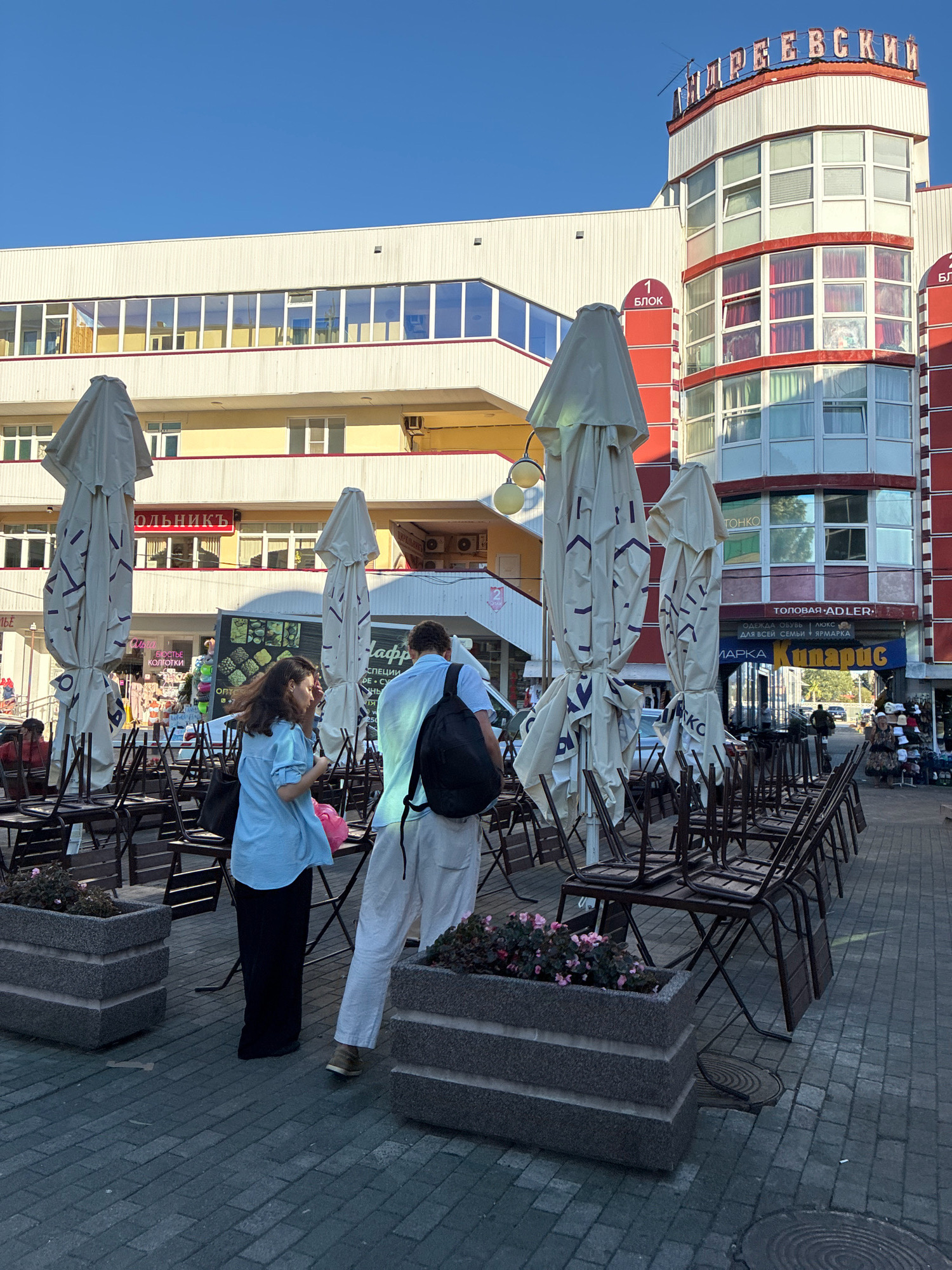 A medium shot captures an outdoor cafe with covered umbrellas and stacked chairs under a bright, clear sky. Two individuals, a woman in a light blue shirt and black pants, and a man in a light blue shirt and white pants with a black backpack, stand near the tables. The background features a multi-story building with red and white accents, bearing Cyrillic signage, indicating it is likely a commercial establishment. The street is paved with dark grey brick. The building's architecture and the Cyrillic text suggest a location in Russia, potentially Sochi, Russia. The scene appears to be during daylight hours, with the sun casting shadows.