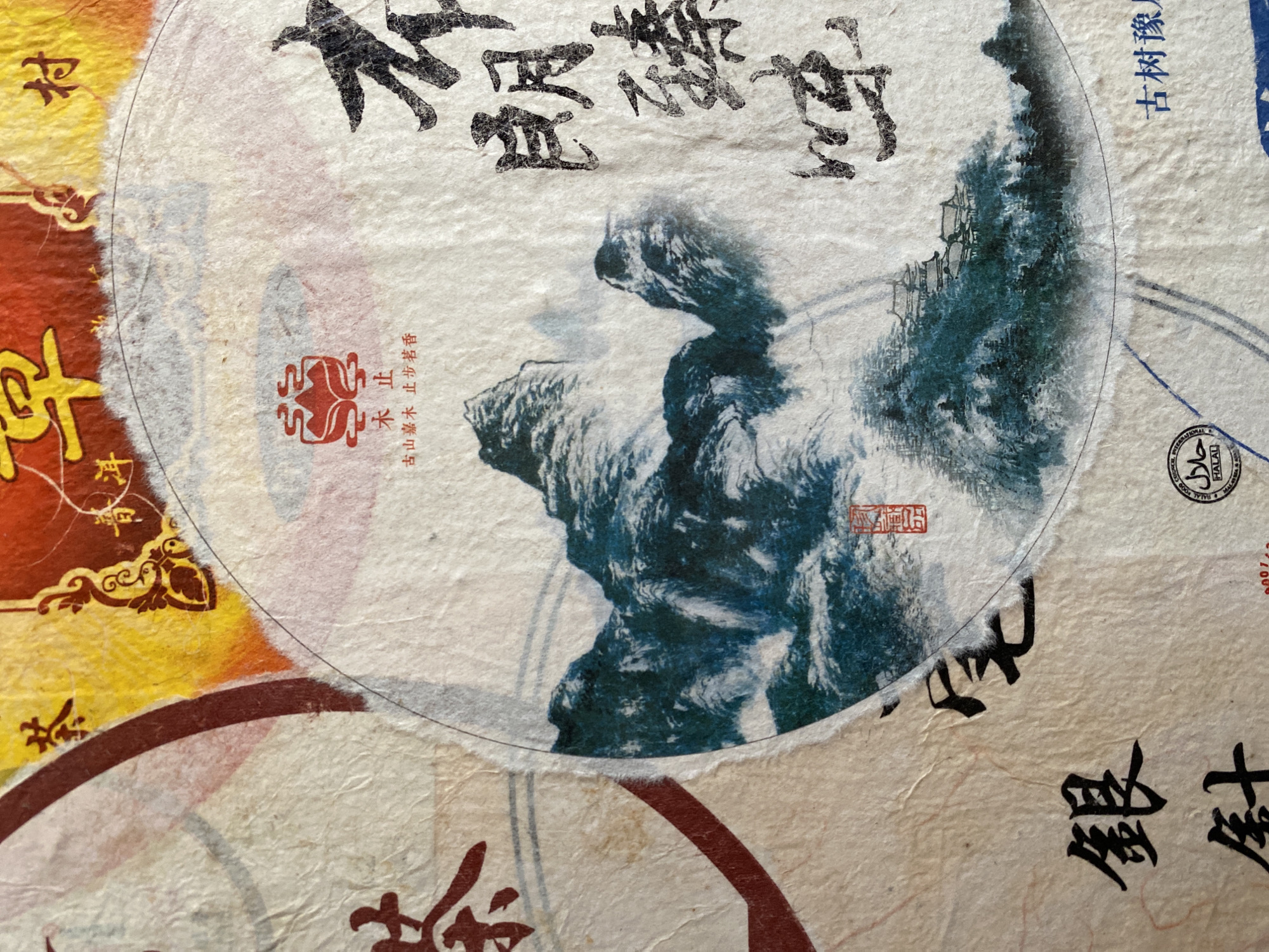 The image shows a close-up of a textured surface, possibly paper, with several overlaid elements. The dominant element is a circle containing an image of a mountain landscape rendered in blue and white tones, reminiscent of traditional Chinese ink painting. Chinese characters are visible above the mountain image, along with a red seal. Other portions of the surface display additional Chinese characters and designs in red, yellow, and black. A Halal certification logo is also visible.