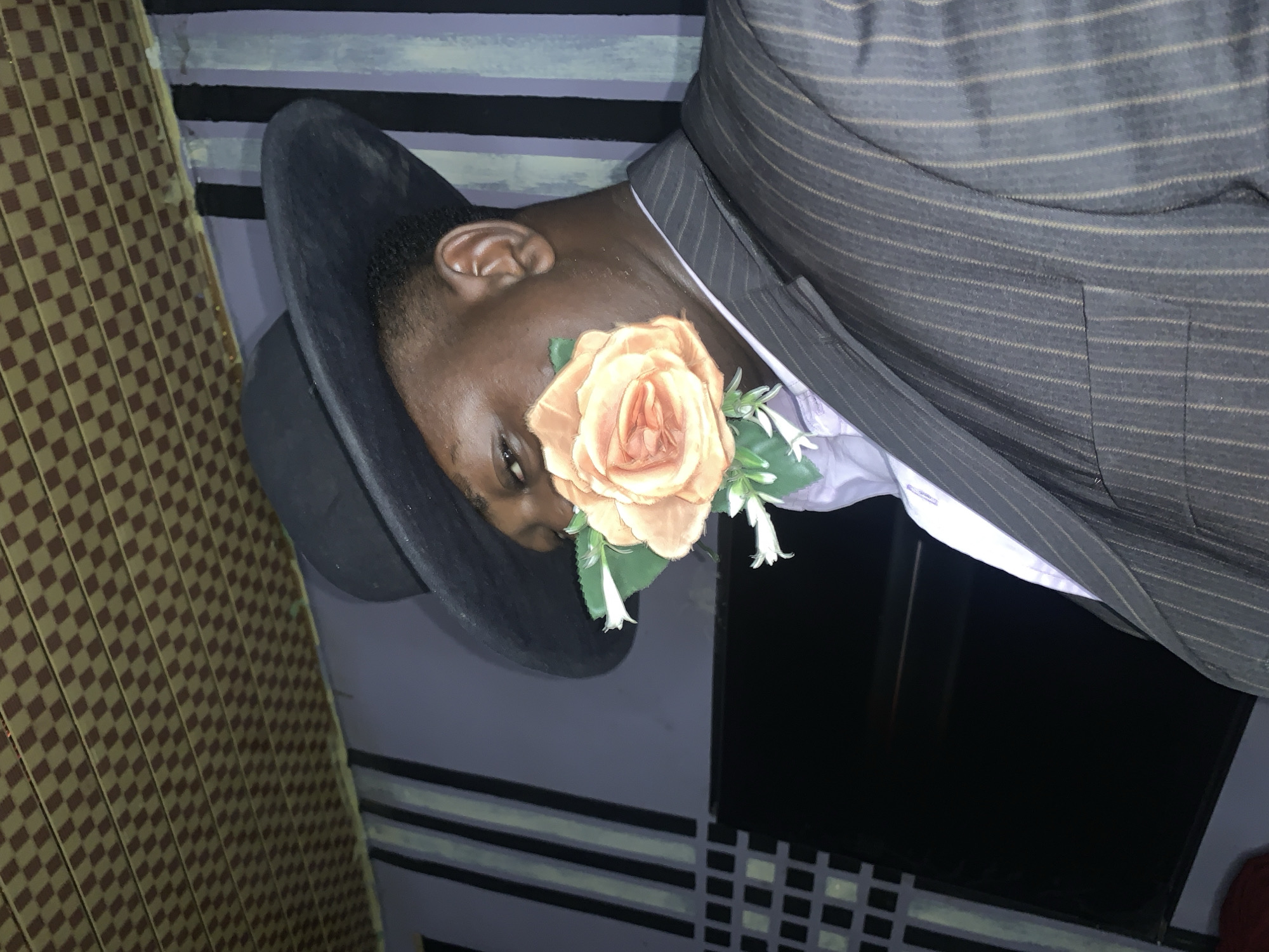 This close-up, rotated image, taken in Akwanga, Nigeria, features an individual, likely a man, from the chest up. The subject has dark skin and is looking downwards, with only the upper part of his face (forehead, eye, ear) clearly visible. He is wearing a black fedora-style hat.
A large, artificial-looking, peach-colored rose, accompanied by smaller white flowers and green leaves, is prominently pinned to the lapel of his suit jacket, situated near his visible eye. The subject is dressed in a dark grey or black pinstriped suit jacket over a white collared shirt that appears to have subtle purple detailing.
The background on the left features a patterned wall or screen with a light brown and dark brown checkerboard design. The central and right portions of the background show a wall with horizontal stripes in varying shades of grey/purple and black, interspersed with some lighter, possibly reflective, horizontal lines. A dark, rectangular area, which could be a screen or a window, is visible in the lower right section of the image.
The image captures the subject in a posed manner, suggesting a formal event, a celebratory occasion, or a stylish portrait. There is no visible text in the image.