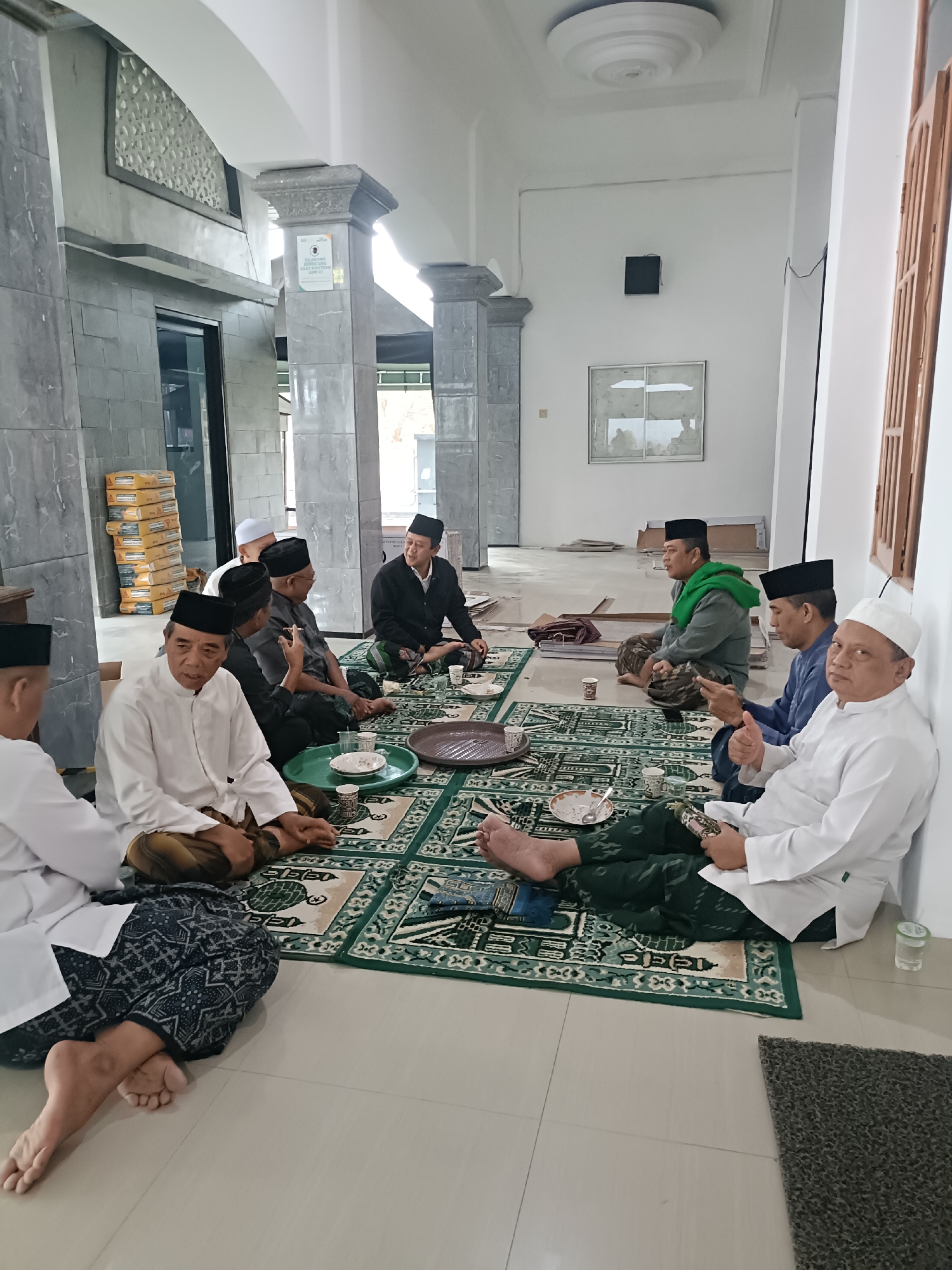 A group of eight men, dressed in traditional Islamic attire, are seated on prayer mats on the floor of what appears to be a mosque or a similar religious building. The men are engaged in conversation, with some holding cups and others gesturing. The environment is sparsely furnished, with stacks of yellow boxes visible in the background, possibly indicating ongoing construction or storage. The floor is tiled, and the walls are a neutral color. One man is wearing a white skullcap and a white tunic, while others are wearing black skullcaps and various colored robes. The overall impression is of a casual gathering or meeting. While the specific location in Kediri City, Indonesia, cannot be definitively determined from the image alone, the context suggests a public or communal space within the city.