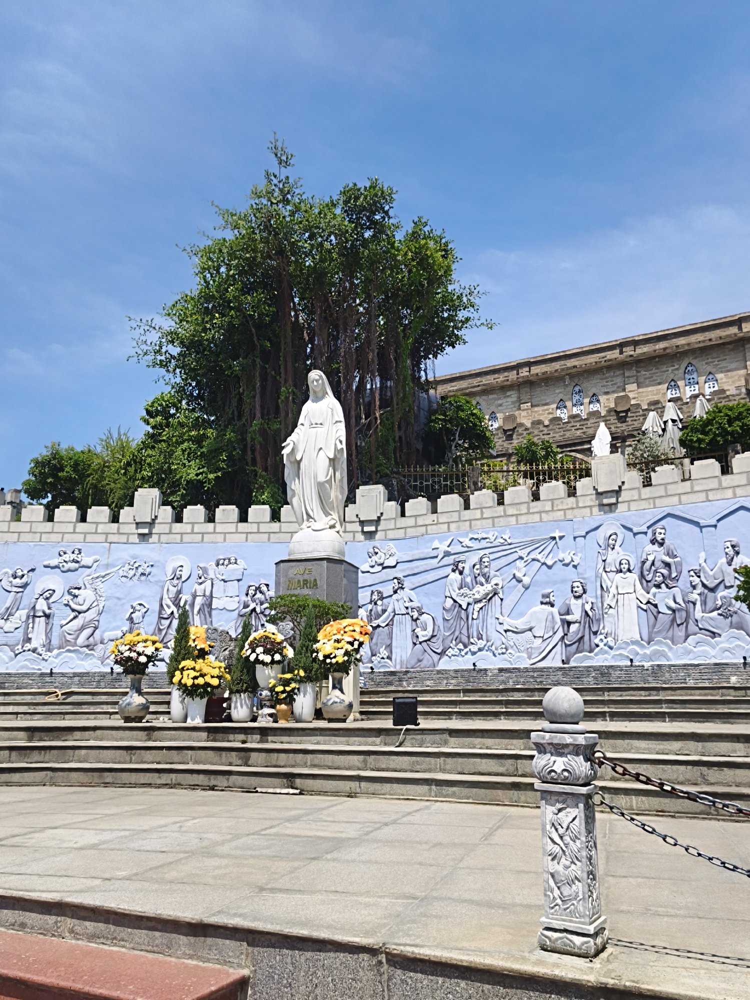 The image depicts a religious monument in Nha Trang Ward, Vietnam. In the foreground, there are stone steps leading up to a plaza. A decorative stone post with a chain fence is visible on the right. Behind the steps, a large blue wall features a detailed relief sculpture depicting various religious figures and scenes. In the center of the relief, a statue of Jesus with a crown of thorns is prominently displayed, with two figures on either side. To the left of the Jesus sculpture, there are figures of angels and other individuals. The wall is topped with a crenellated parapet. A white statue of Mary stands on a pedestal in front of the wall, to the right of the Jesus sculpture. The inscription "AVE MARIA" is visible on the pedestal of the Mary statue.
Behind the wall, a large, mature tree with visible aerial roots dominates the upper left portion of the image. In the background, a stone building with arched windows and a tiered facade is visible. The sky above is a clear, bright blue. Several vases of flowers are arranged on a lower level in front of the relief wall, adding color to the scene. The overall atmosphere suggests a sacred and serene location.