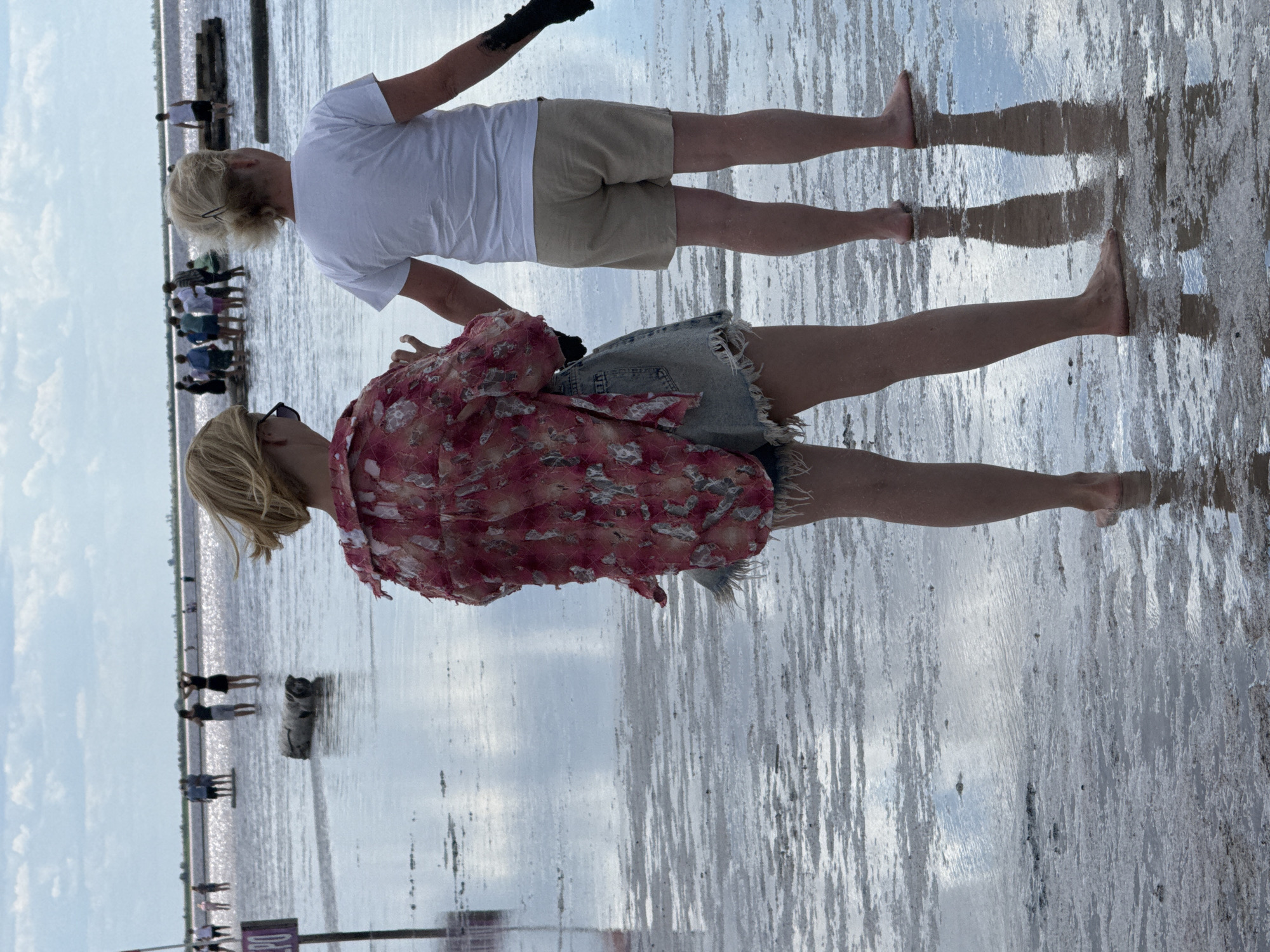 This is a medium shot of two women standing in shallow water at a salt lake in Bursol, Russia. The woman in the foreground is facing away from the camera, wearing a red and white patterned shirt and denim shorts. She has blonde hair and is barefoot. The woman in the background is also facing away from the camera, wearing a white t-shirt and beige shorts. She has blonde hair and is barefoot. Both women appear to be holding hands or reaching towards each other. The water is shallow and reflects the cloudy sky. In the background, other people can be seen standing in the water and on wooden platforms. The ground is covered in salt deposits.
