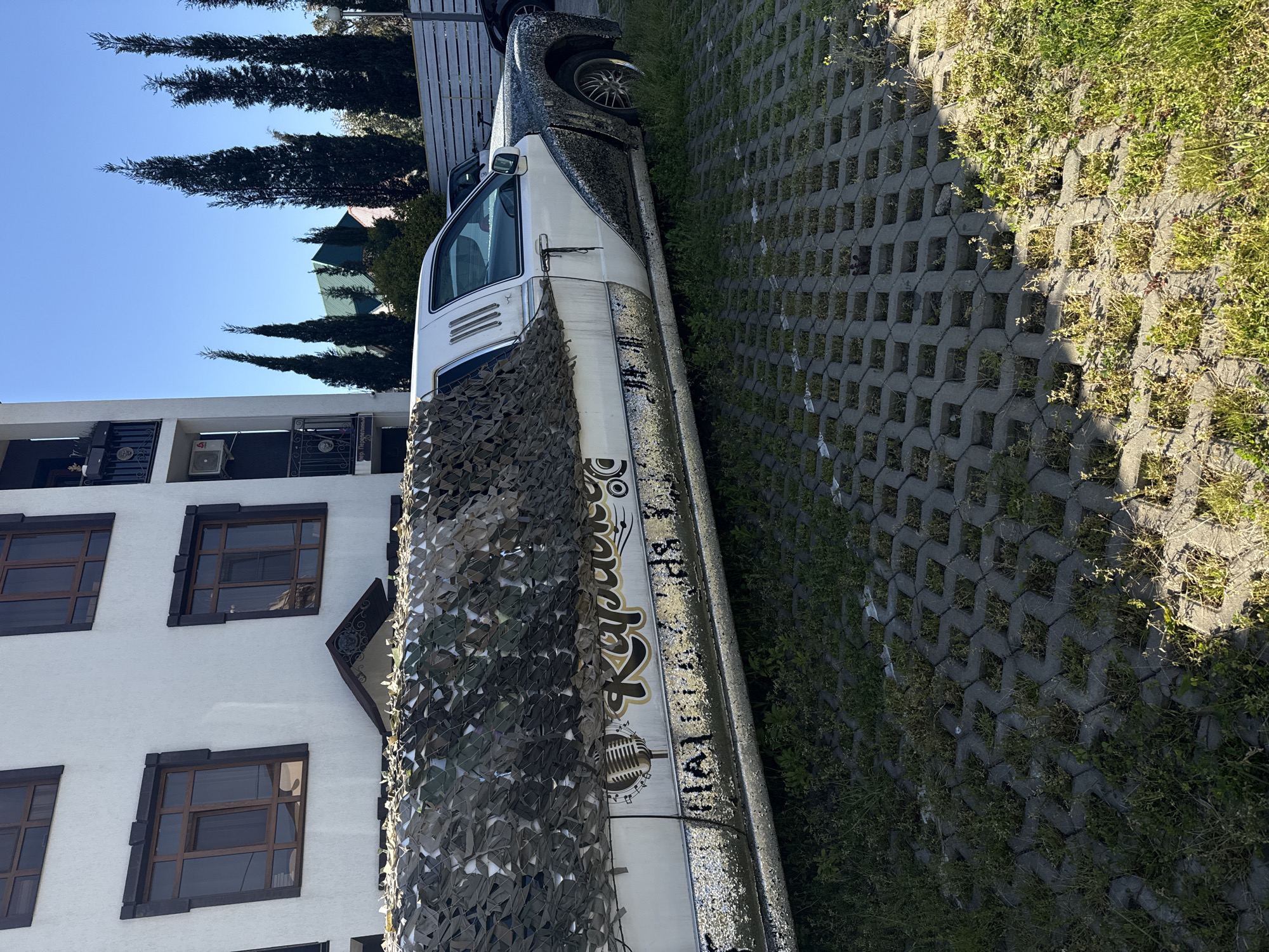 The image displays an outdoor scene in Sochi, Russia, featuring a customized vehicle parked on patterned pavers next to a white multi-story building, beneath a clear blue sky. Bright sunlight illuminates the scene, indicating daytime.
The central subject is a long, highly modified vehicle, resembling a limousine or a large van, with a white lower body and a dark, possibly glitter-covered, upper section. A camouflage-patterned net is draped over the front passenger side. On the white section of the vehicle, a prominent gold and white stylized logo reads "Караоке" (Karaoke), accompanied by a microphone icon. Below this, the text "Поехали!" (Let's go!) is visible. A silver multi-spoke wheel is seen on the front axle.
To the left of the vehicle stands a modern white building featuring dark-framed rectangular windows. Balconies with ornate black metal railings are present, and an air conditioning unit is mounted on one. A small sign visible on a balcony railing identifies the location as "ул. Подгорная" (Podgornaya Street).
The ground surface is covered with grey interlocking concrete pavers, arranged in a grid-like pattern, with green grass and weeds growing through the gaps. In the background, behind the building and vehicle, several tall, slender, dark green cypress trees stand against the vibrant, cloudless blue sky. No people are visible in the frame.
