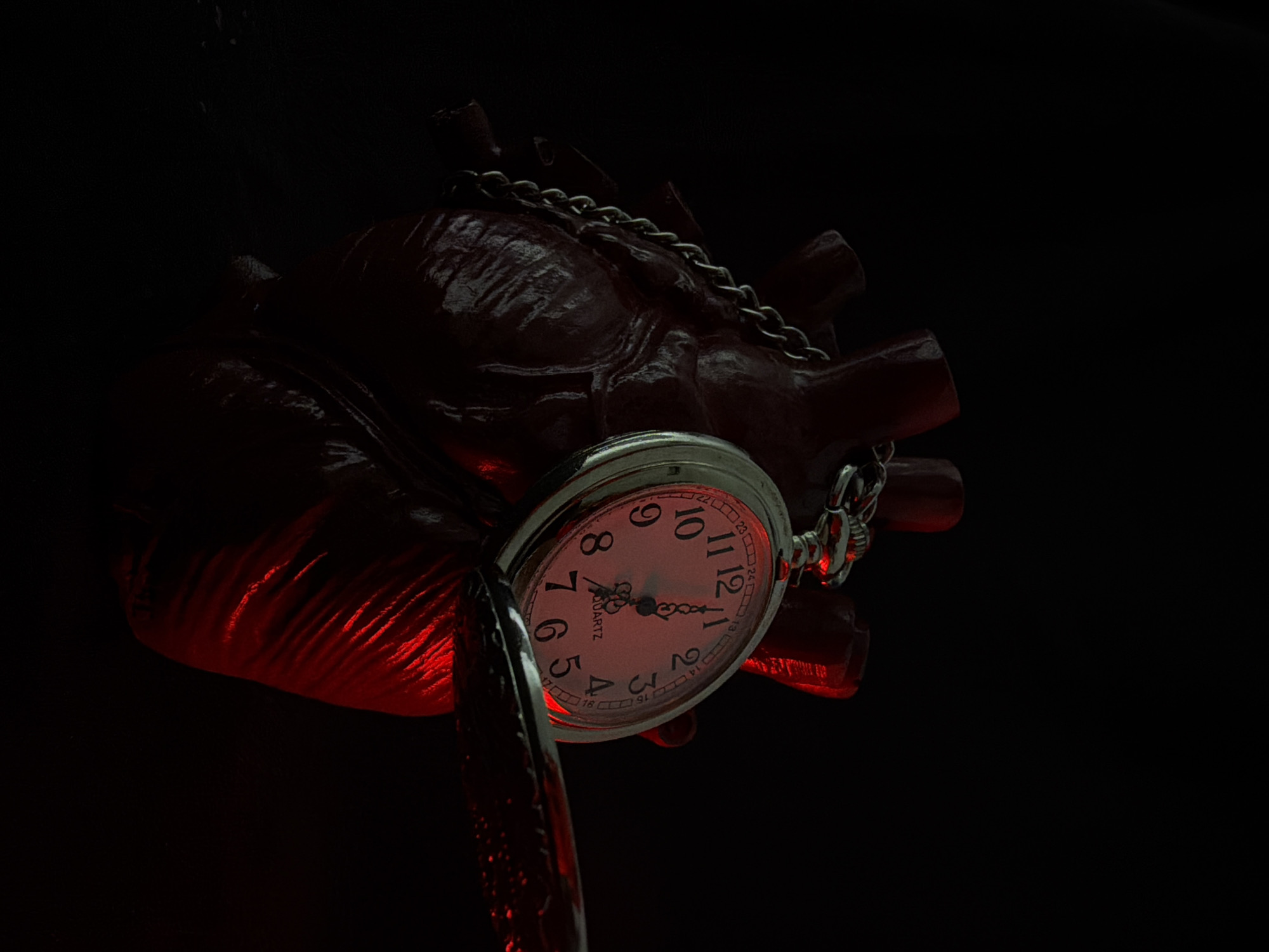 A close-up, dimly lit shot focuses on a red, heart-shaped object, possibly a sculpture or prop, against a black background. A silver pocket watch is attached to the heart by a chain, with the watch face open. The watch has Roman numerals and a white face, with the time appearing to be just past 11:00. Red light illuminates the heart from below, casting sharp highlights and shadows, particularly on the ribbed surface of the object. The overall mood is dramatic and slightly unsettling, with the juxtaposition of the anatomical heart and the timekeeping device suggesting themes of mortality, urgency, or the passage of time in relation to life. The scene is devoid of any discernible environmental or location cues, and no people are visible. The only visible text is "QUARTZ" printed on the watch face.