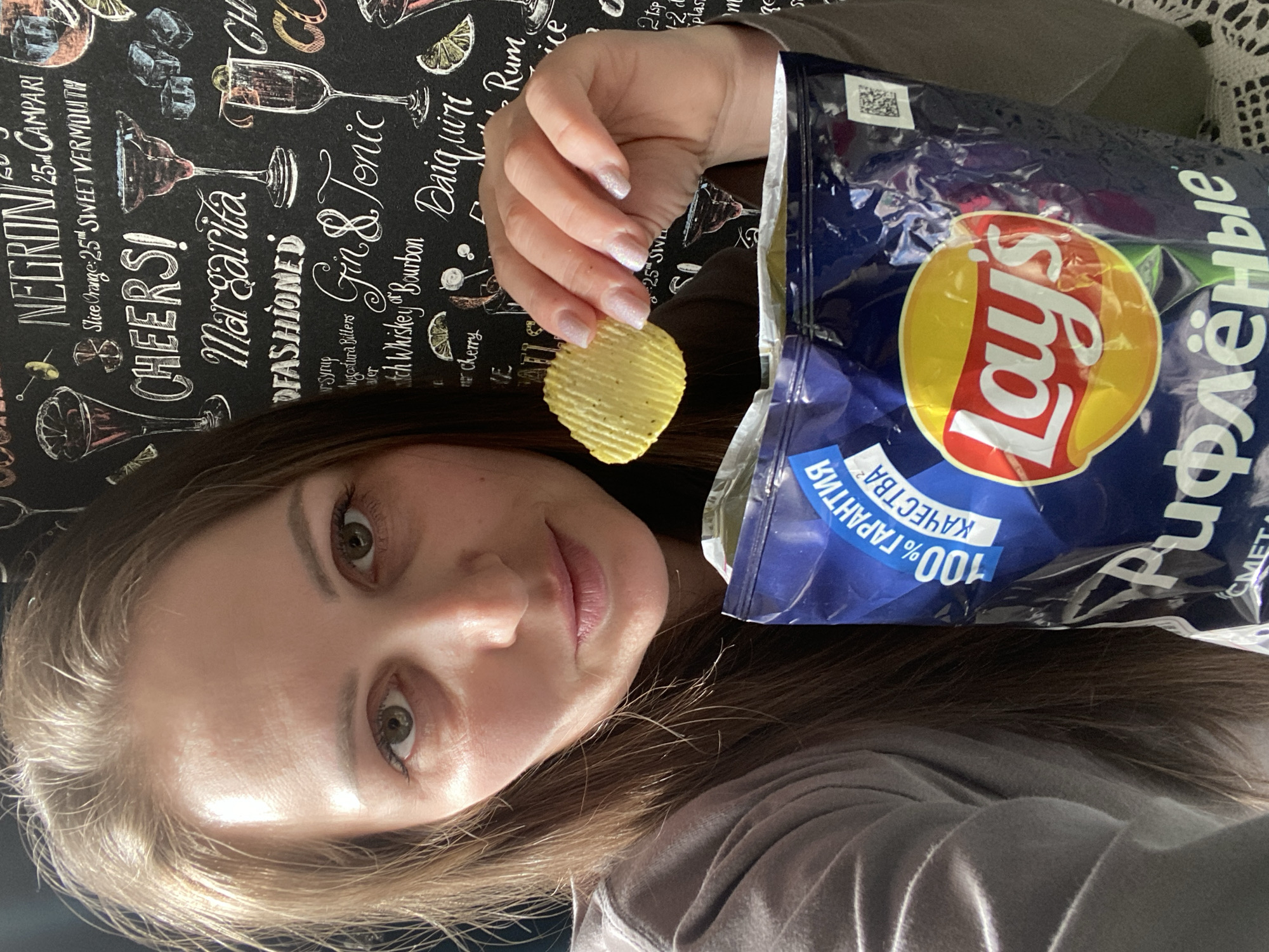 A woman with light brown hair is taking a selfie while holding a ridged potato chip in her right hand. She is looking at the camera with a slight smile. Behind the chip, she holds open a blue bag of Lays "Rifleniye" (ridged) chips. The bag has Russian writing and a "100% quality guarantee." Behind the woman is a black tablecloth with white drawings and names of alcoholic drinks, such as "Negroni," "Margarita," and "Gin & Tonic." The scene likely takes place indoors. There is no specific information to indicate the location is in Markovskii, Russia.