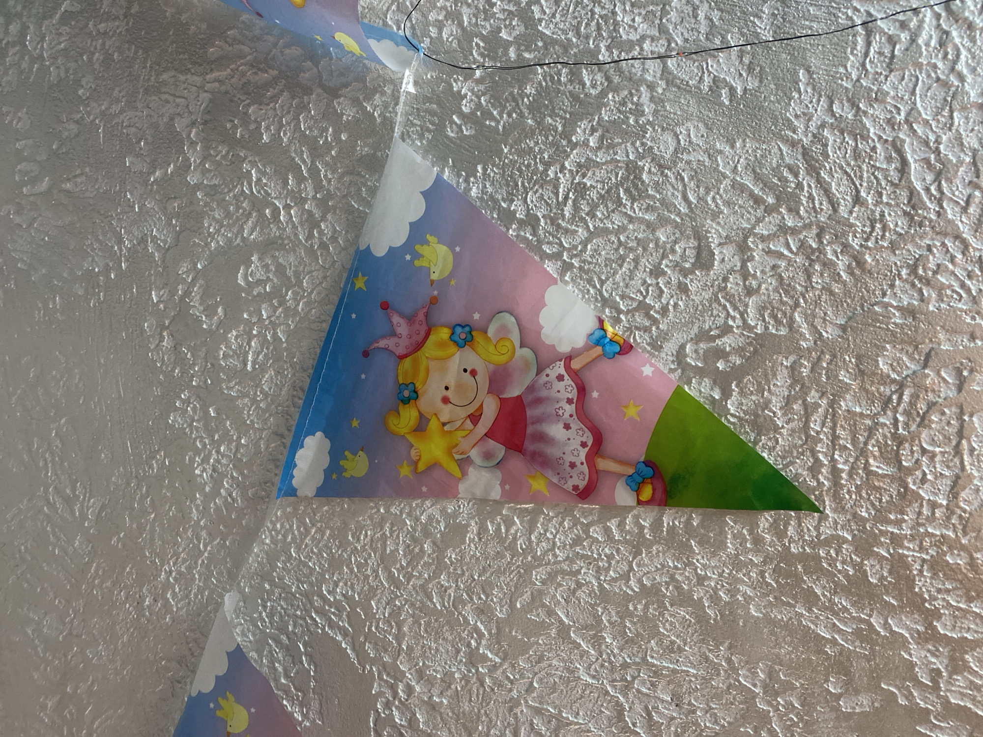 A close-up, eye-level shot shows a decorative garland of triangular flags hanging on a textured, white ceiling. The garland features an illustration of a fairy with blonde hair, wearing a pink dress and a crown, holding a yellow star. The background of the flag is a gradient of blue and pink with clouds and yellow birds. The ceiling has a rough, stucco-like texture. There is a thin black wire visible on the right side of the image, also attached to the ceiling. This scene is located in Kryvyi Rih, Ukraine.