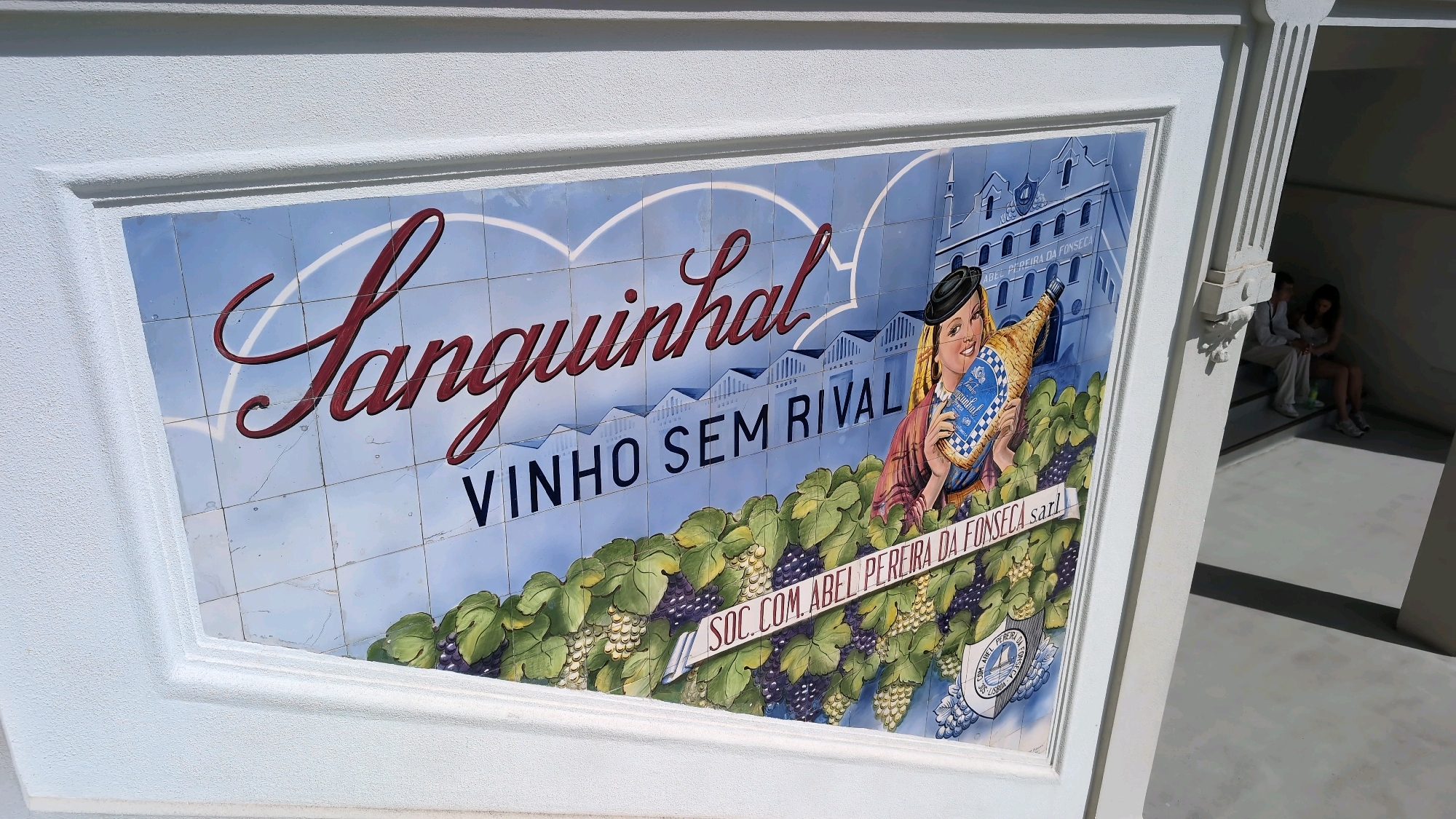 The image shows a large ceramic tile mural on the exterior of a building, likely in Porto, Portugal, given the context of Portuguese wine. The mural depicts a scene related to wine production, featuring the brand name "Sanguinhal" in large, stylized red lettering. Below "Sanguinhal," the phrase "VINHO SEM RIVAL" (Wine without rival) is written.
The mural also includes a drawing of a smiling woman wearing a black hat and holding a bottle of Sanguinhal wine, with a label that reads "Vinho Sanguinhal." Behind her, there are illustrations of a building, possibly a winery, with multiple windows and a tower, and a row of smaller buildings. The foreground of the mural is filled with depictions of grapevines, including bunches of dark purple and light green grapes.
A banner runs across the bottom of the mural, above the grapevines, with text that appears to read "SOC. COM. ABEL PEREIRA DA FONSECA sarl," indicating the company associated with the wine. A circular emblem or crest is also visible on the right side of the mural.
To the right of the mural, there is an architectural element of the building with angular white trim and shadow casting, leading into a shadowed entrance or alcove. Two people, a male and a female, are seated on steps within this alcove. The man is wearing a white shirt and the woman is wearing a light-colored top. They appear to be resting or waiting. The lighting suggests it is daytime, with sunlight casting shadows. The overall scene conveys a sense of heritage and tradition associated with Portuguese wine.