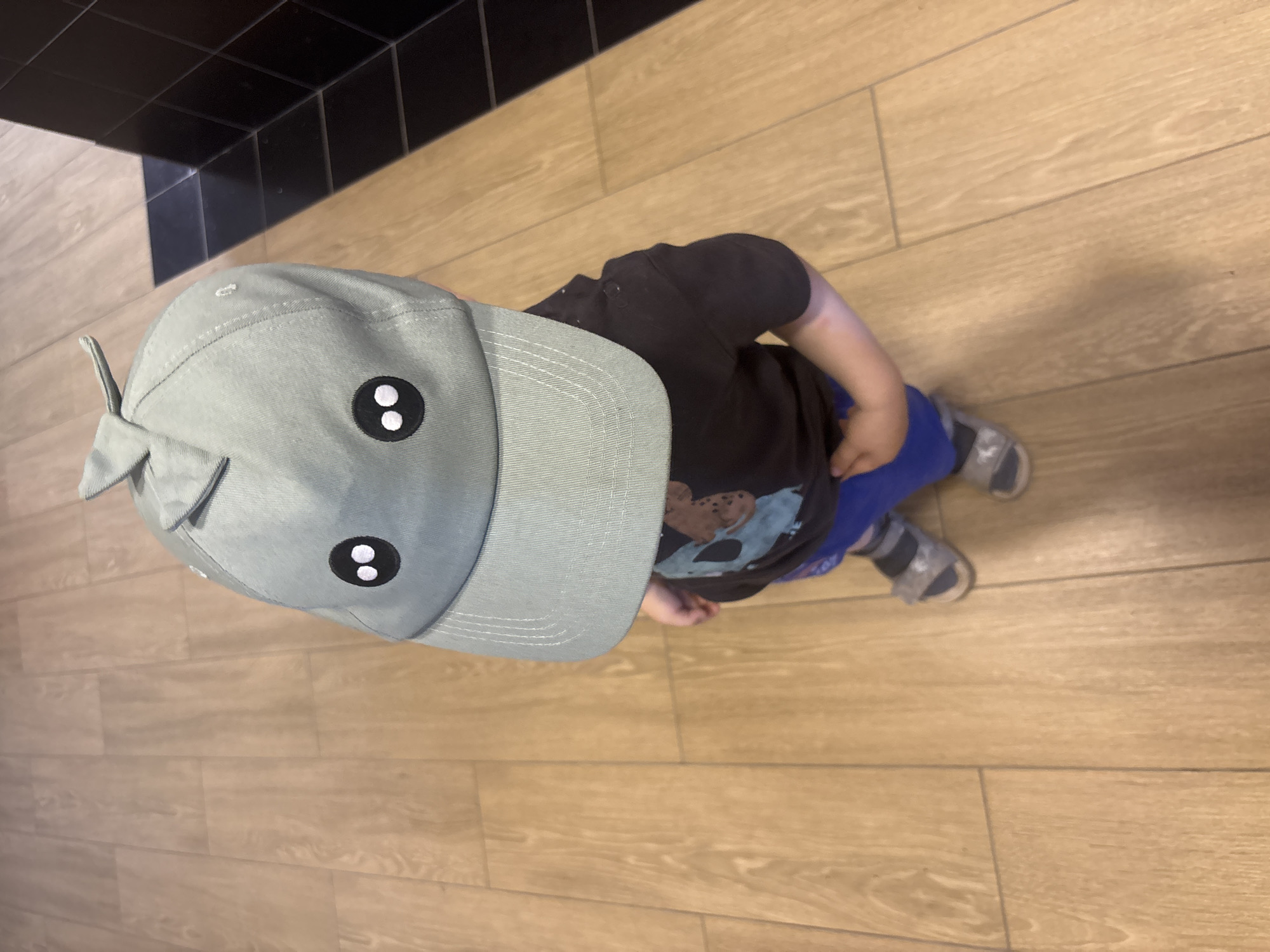 A small child is standing on a light-colored tiled floor. The child is wearing a grey hat with eyes, a dark t-shirt, blue pants, and grey sandals. The floor appears to be indoors and the tiled pattern simulates a wood-like texture. A black tiled area is partially visible in the upper left corner of the frame. The child is viewed from directly above.