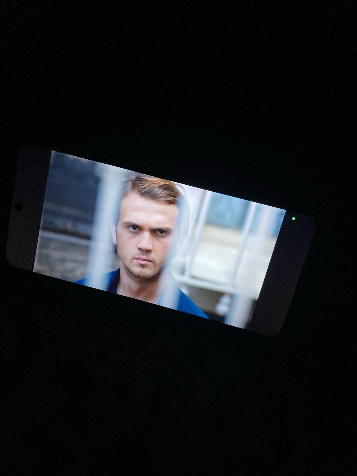 The image displays a smartphone screen angled from the top right to the bottom left. The screen shows a close-up, slightly blurred portrait of a young Caucasian man with short, light brown hair and a serious expression. He is wearing a blue collared shirt. The background of the image on the screen appears to be an outdoor setting with muted colors and some vertical lines, possibly indicating a fence or blinds. A small green light is visible on the right edge of the smartphone screen. The surrounding environment is completely black, suggesting the photo was taken in a dark room or at night. The location in the city of Ramenskoye, Russia, is not discernible from the image content.